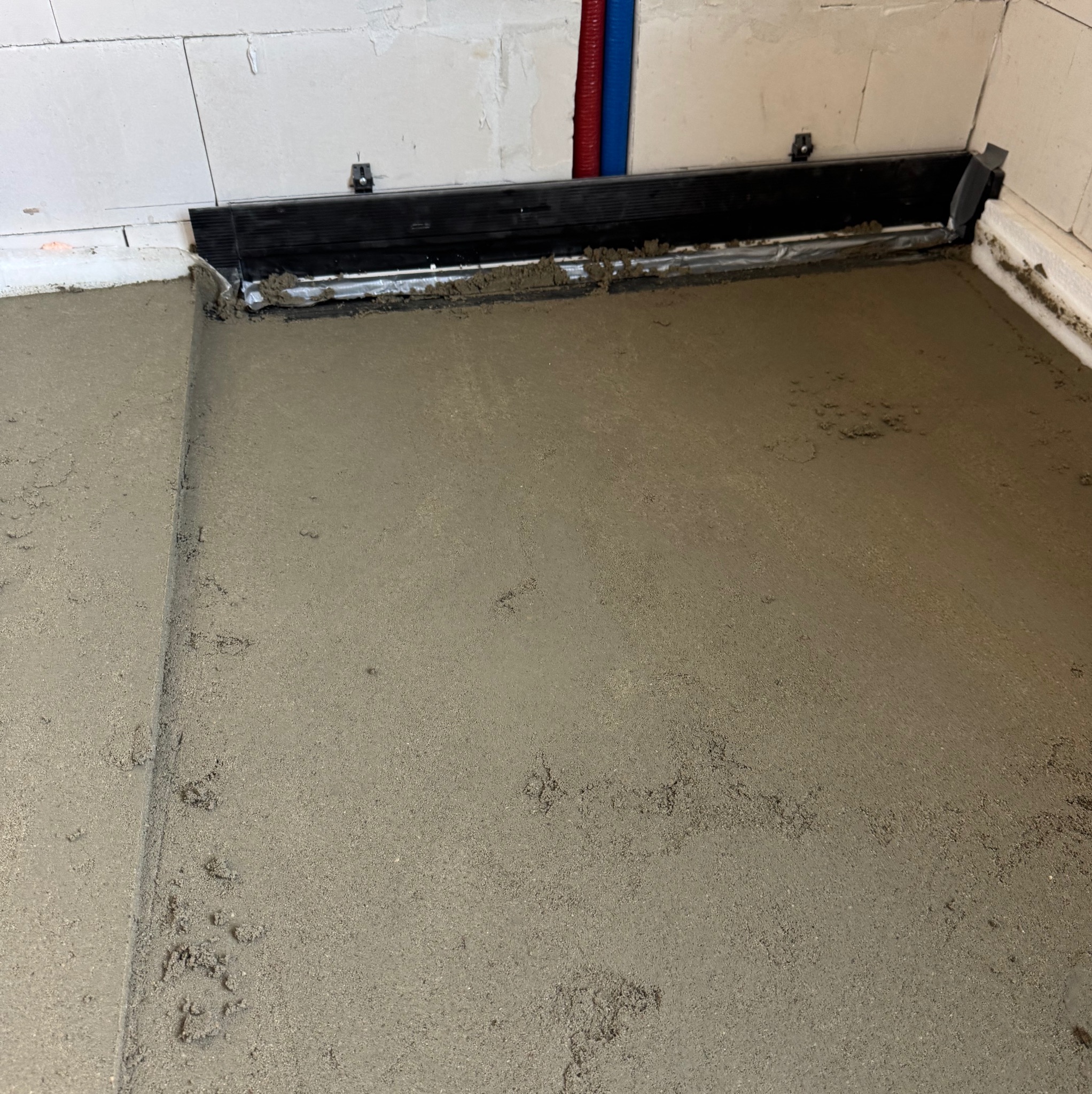 Świeżo wylana, szara posadzka jastrychowa z widocznymi listwami dylatacyjnymi przy ścianach z białych bloczków. Czerwona i niebieska rurka w tle.