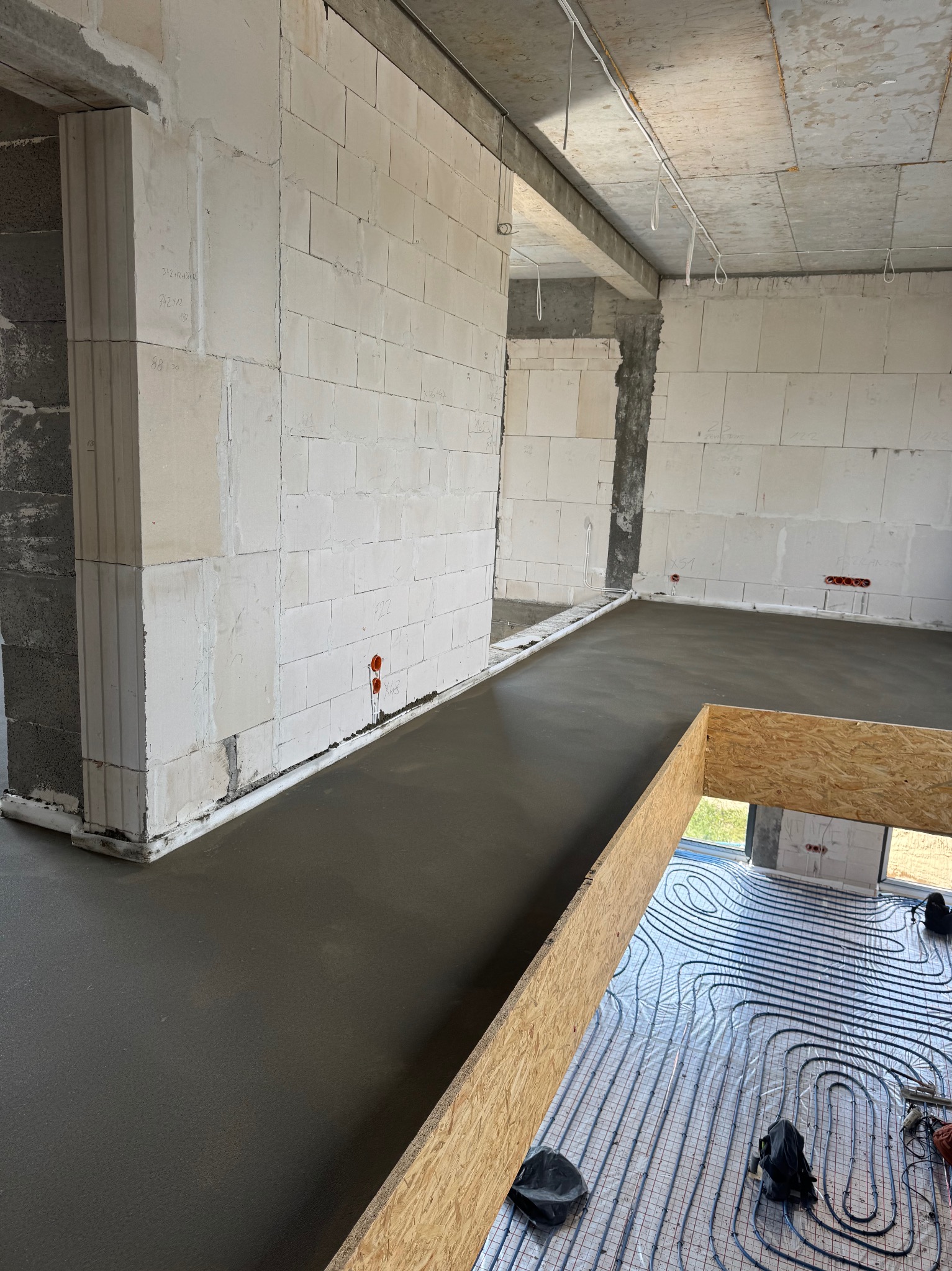 Wnętrze w trakcie wylewania posadzki jastrychowej. Widoczne ogrzewanie podłogowe i surowe ściany z betonu komórkowego. Częściowo zalana podłoga.