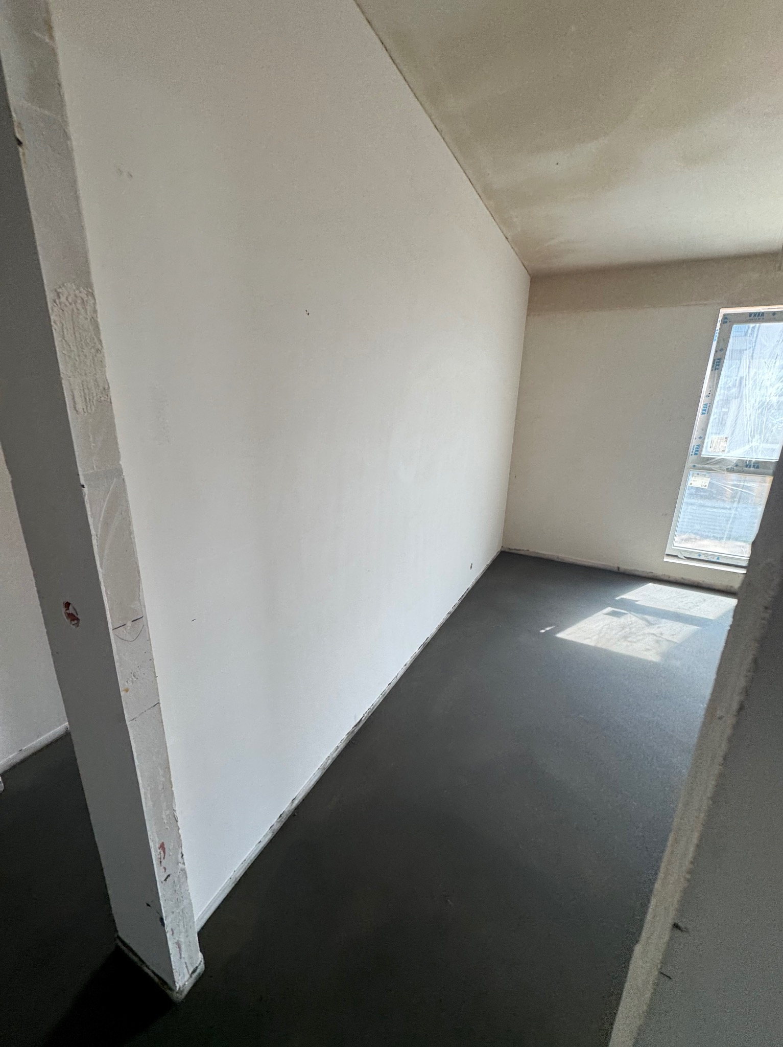 Wnętrze z wylaną, ciemną posadzką jastrychową, ściany przygotowane do malowania, okno z widoczną folią ochronną. Surowy stan wykończenia.