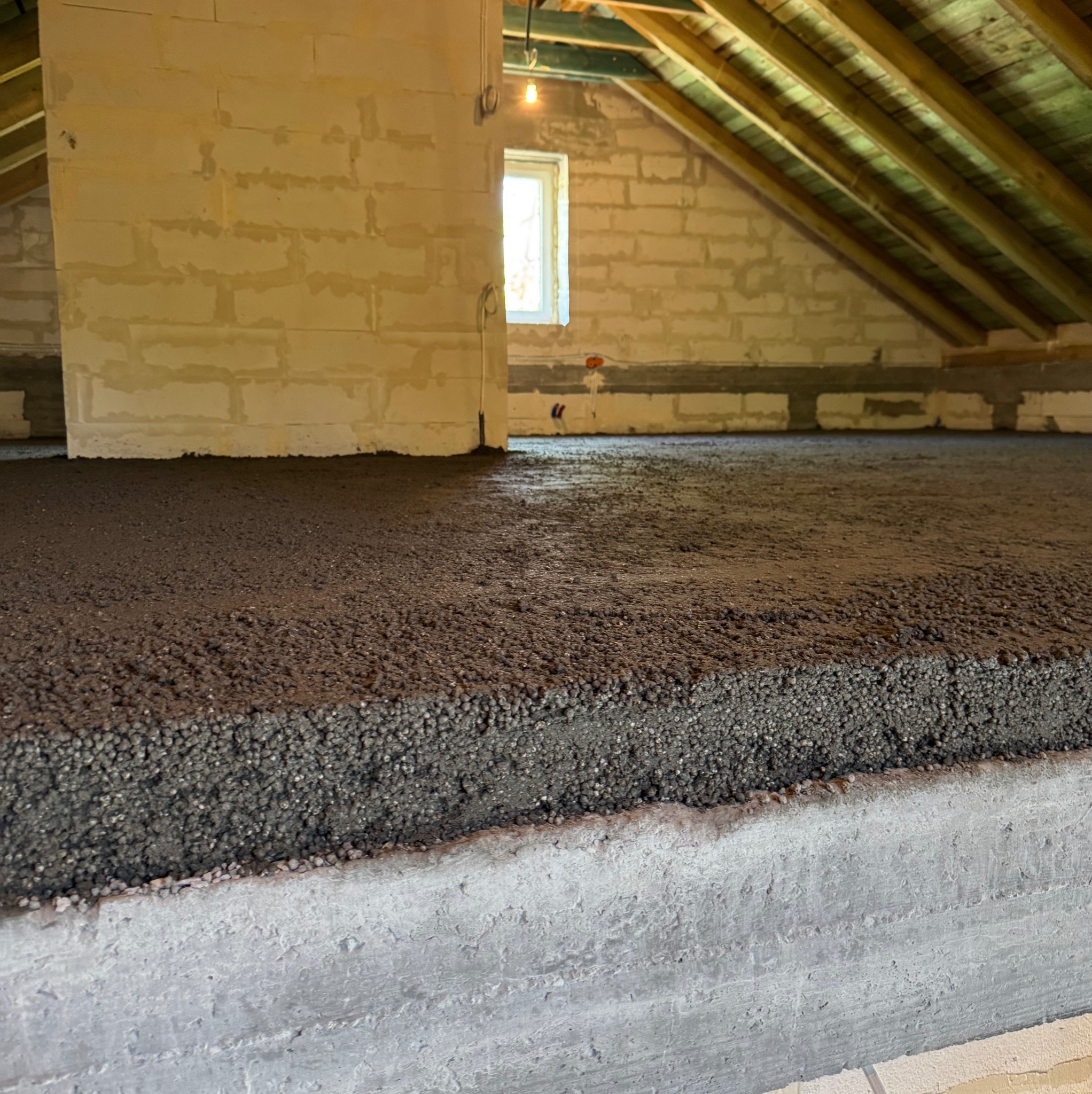 Szary jastrych styrobetonowy na poddaszu, widok z bliska na strukturę materiału i betonową wylewkę, surowe ściany z bloczków i drewniana konstrukcja dachu.