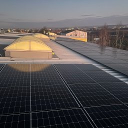 Fujity - Instalacja paneli słonecznych na dachu budynku przemysłowego o zmierzchu, widoczni pracownicy w odblaskowych kamizelkach, w tle zabudowa miejska.