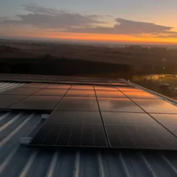 Panele słoneczne na dachu budynku odbijające wschodzące słońce, w tle krajobraz z budynkami i drzewami w porannej mgle.