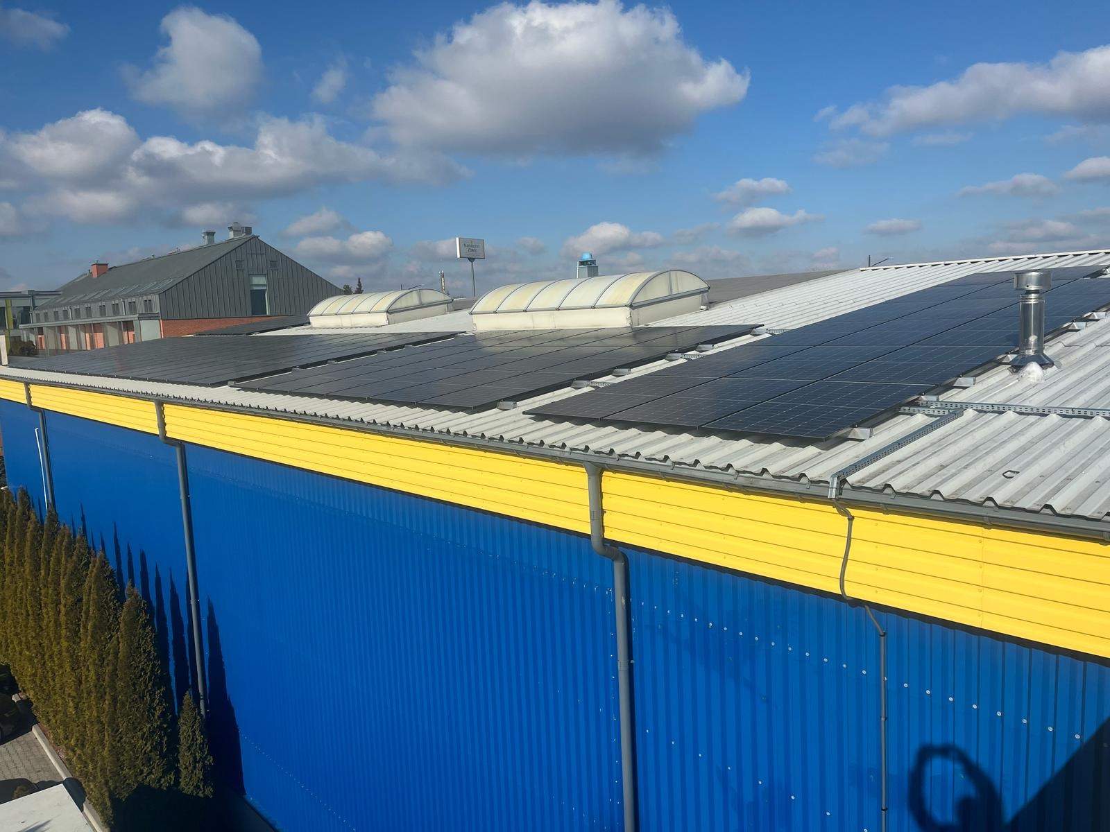 Panele słoneczne na dachu magazynu z niebiesko-żółtą elewacją, widoczne kominki wentylacyjne i błękitne niebo z chmurami.