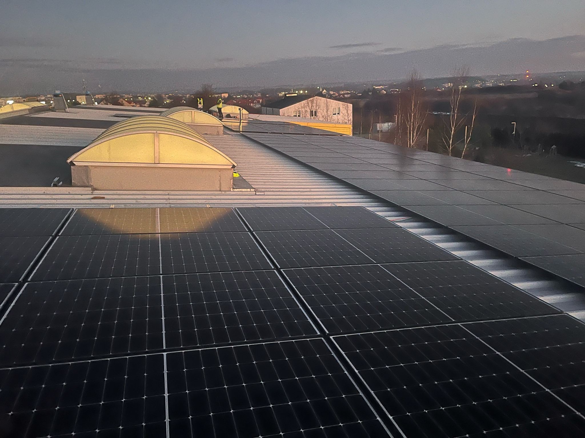 Panele fotowoltaiczne na dachu budynku przemysłowego z świetlikami, w tle widok na miasto o zmierzchu.