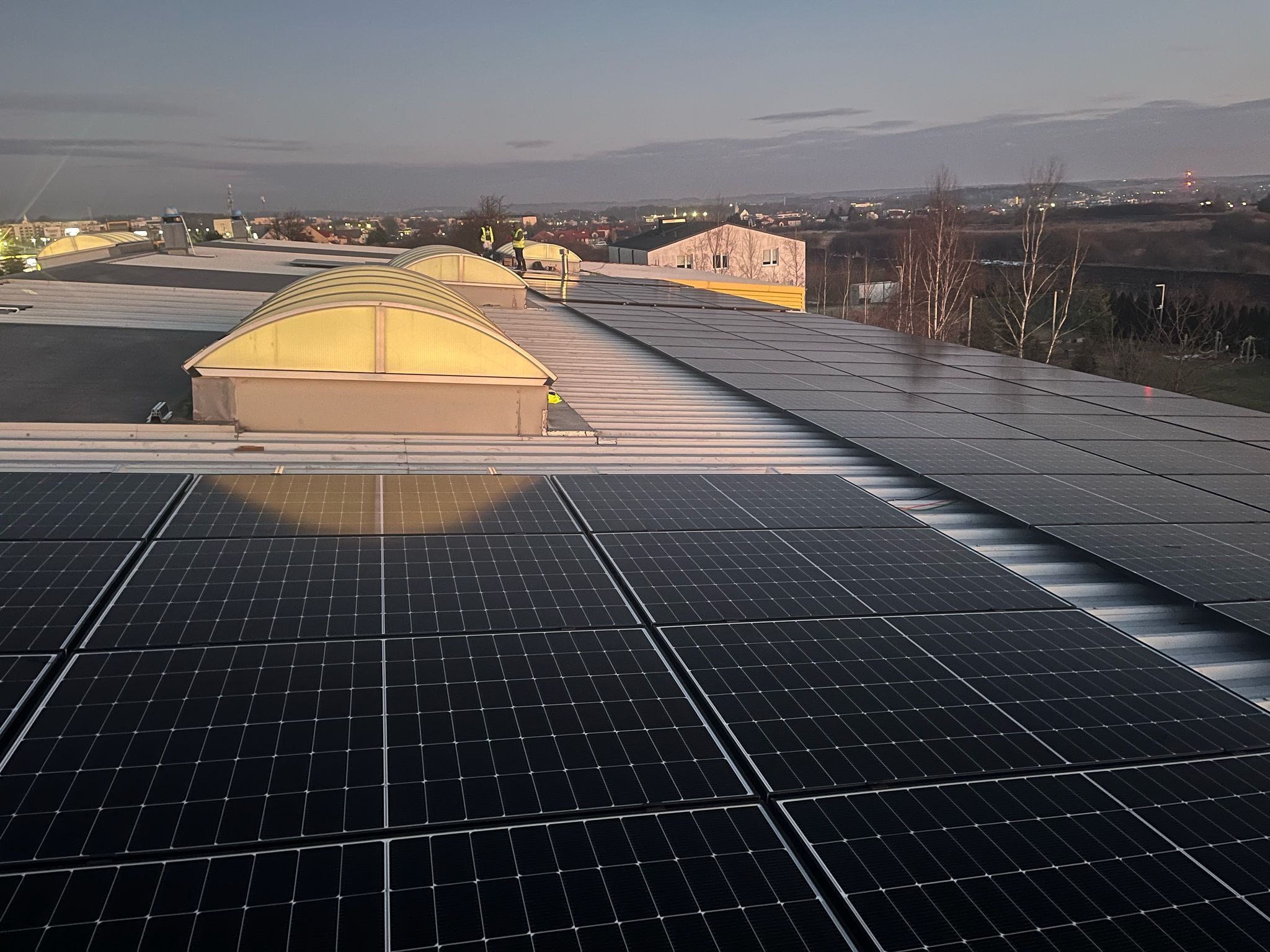 Instalacja paneli słonecznych na dachu budynku przemysłowego o zmierzchu, widoczni pracownicy w odblaskowych kamizelkach, w tle zabudowa miejska.