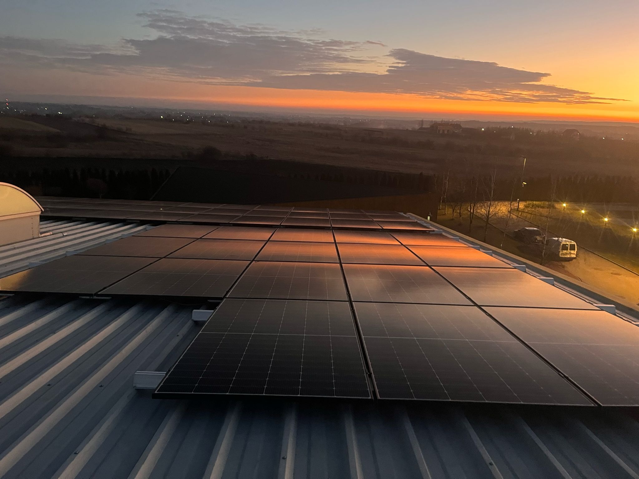 Panele słoneczne na dachu budynku odbijające wschodzące słońce, w tle krajobraz z budynkami i drzewami w porannej mgle.
