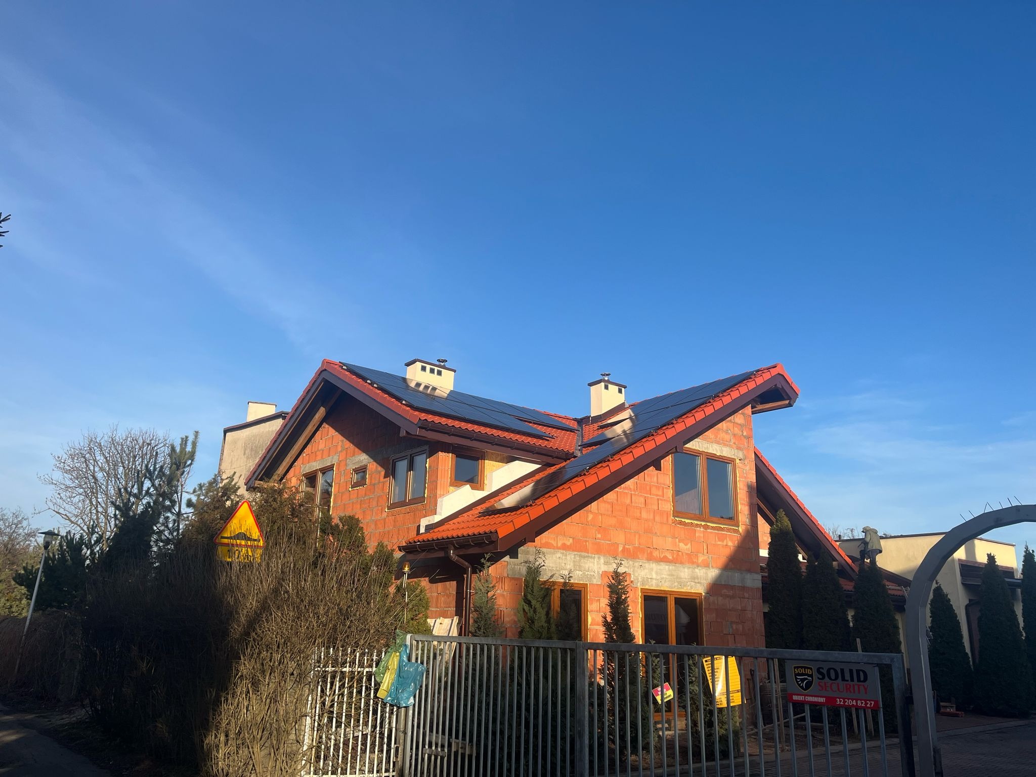 Dom w trakcie budowy z częściowo zamontowanymi panelami fotowoltaicznymi na dachu, widoczne ogrodzenie i tabliczka agencji ochrony.