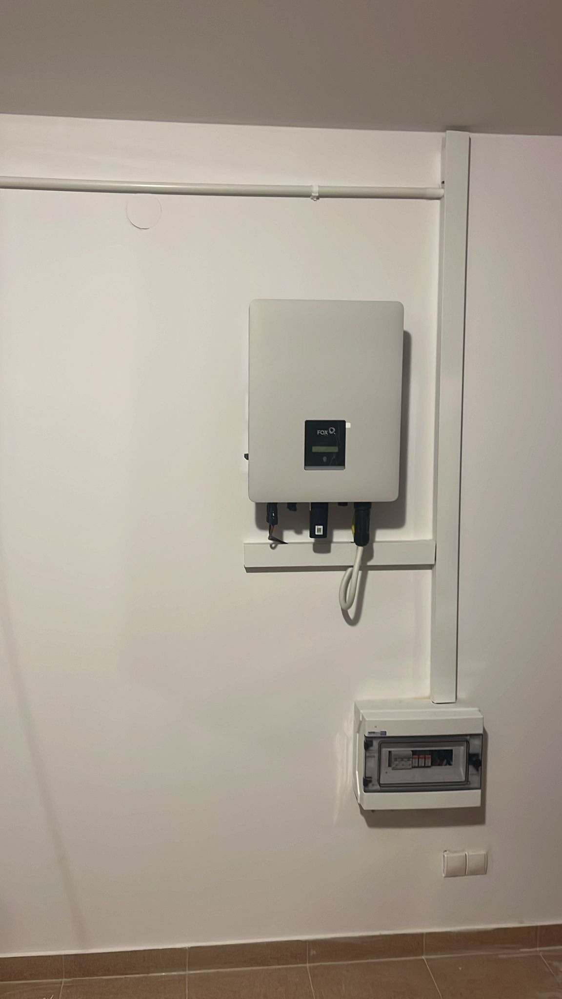 Biały inwerter fotowoltaiczny marki FOX zamontowany na białej ścianie nad skrzynką bezpiecznikową z widocznymi przewodami, z rurą instalacyjną biegnącą wzdłuż ściany.