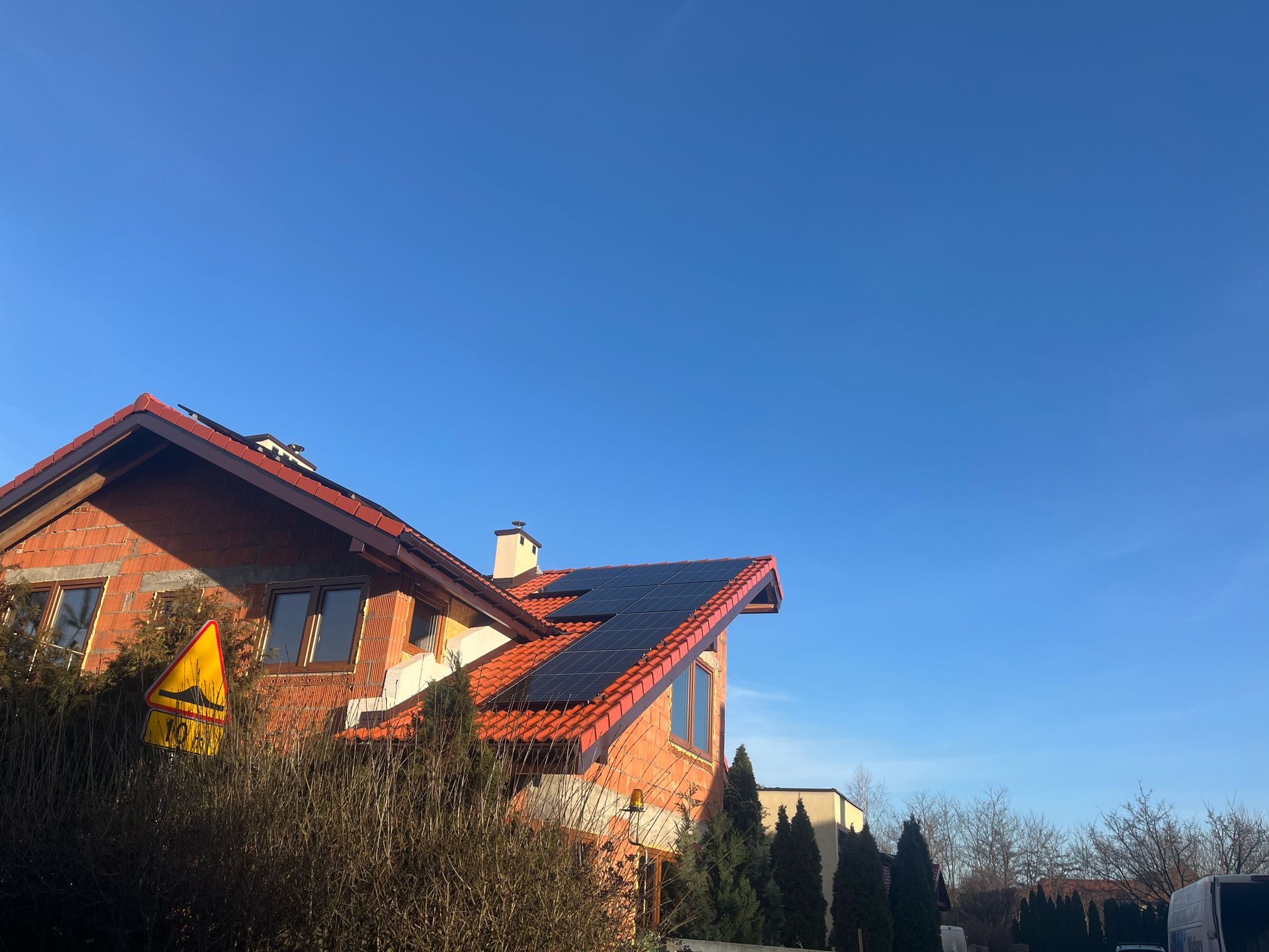 Dom jednorodzinny w trakcie budowy z zamontowanymi panelami fotowoltaicznymi na czerwonym dachu, widoczny znak drogowy ostrzegający o nierównościach.