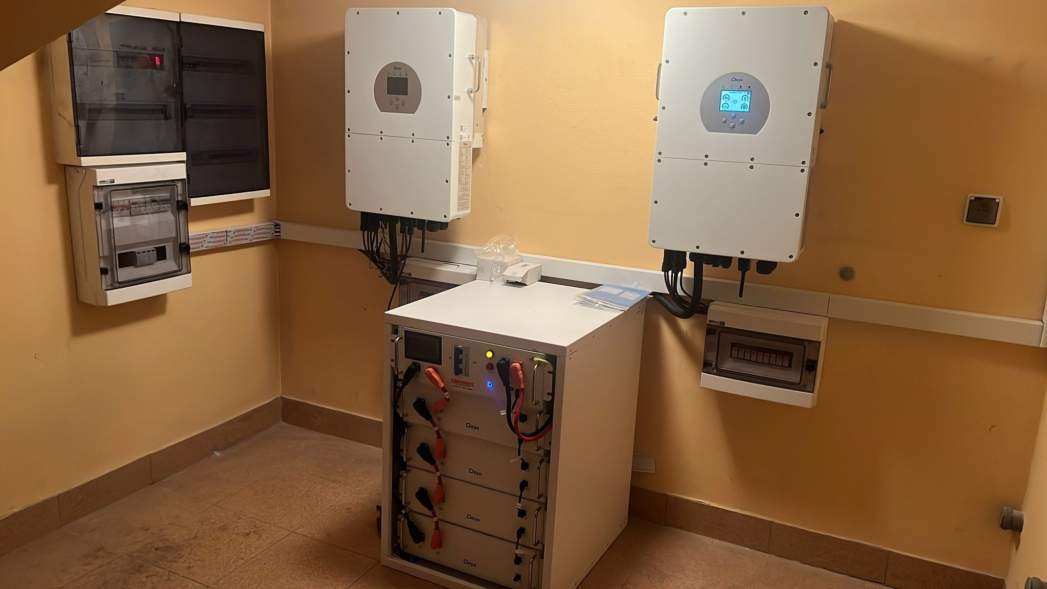 Wewnętrzna instalacja fotowoltaiczna z dwoma inwerterami na ścianie i stojącym modułem baterii magazynu energii, widoczna skrzynka elektryczna i przewody.