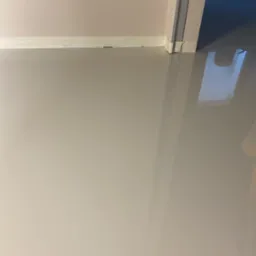 Jasnoszara, gładka posadzka z żywicy, zabezpieczona taśmą malarską przy ścianie i ościeżnicy drzwi, z widocznym odbiciem światła.