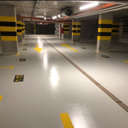 SOBOL - Biała, błyszcząca posadzka w garażu podziemnym z żółtymi oznaczeniami stanowisk, numerami i strzałkami kierunkowymi; kolumny pomalowane w czarno-żółte pasy.