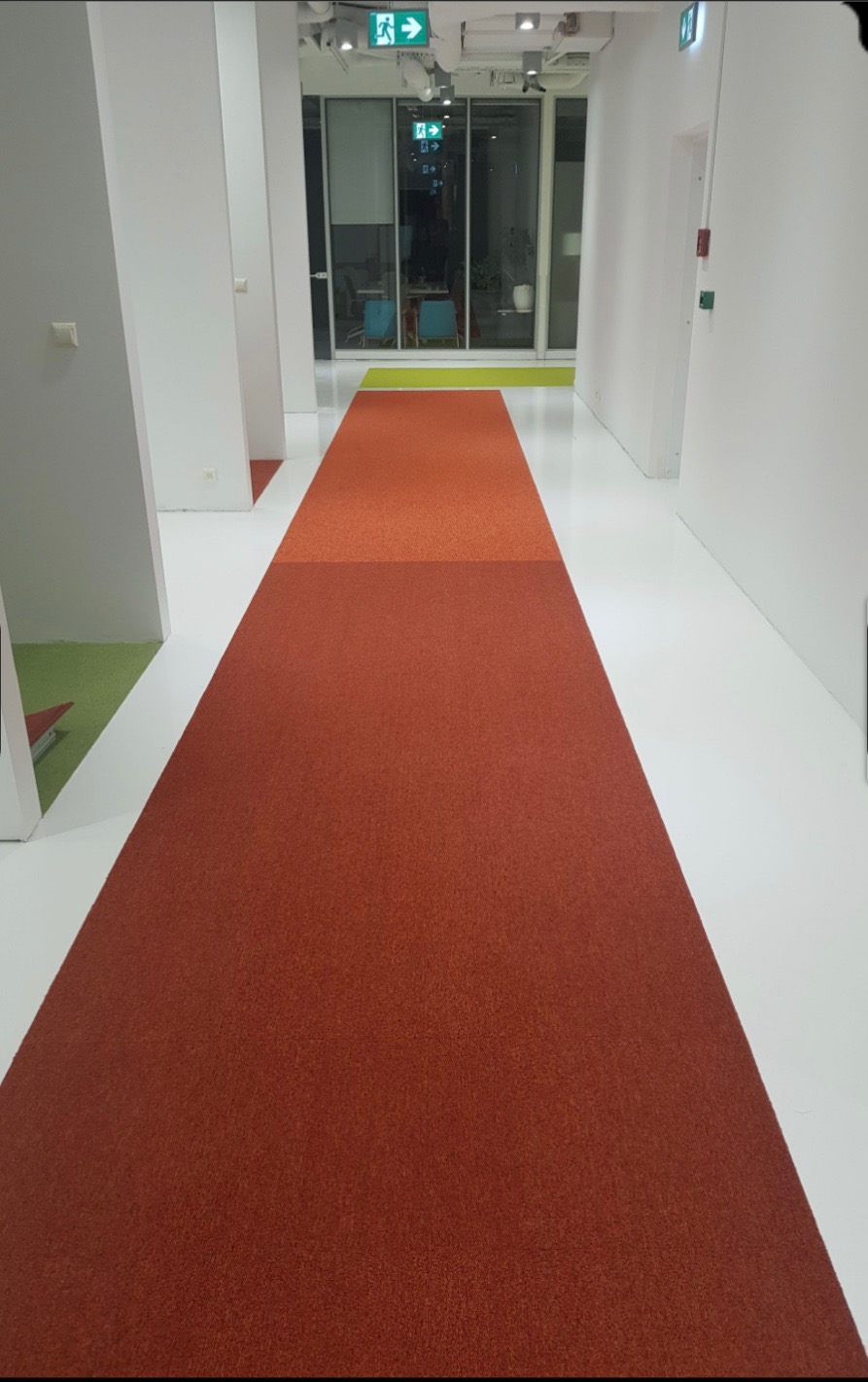 Długi korytarz z pomarańczową, teksturowaną podłogą w odcieniach cegły, przechodzącą w jaśniejszy odcień w oddali, z białymi ścianami i sufitem, oświetlony światłem z okien na końcu korytarza.