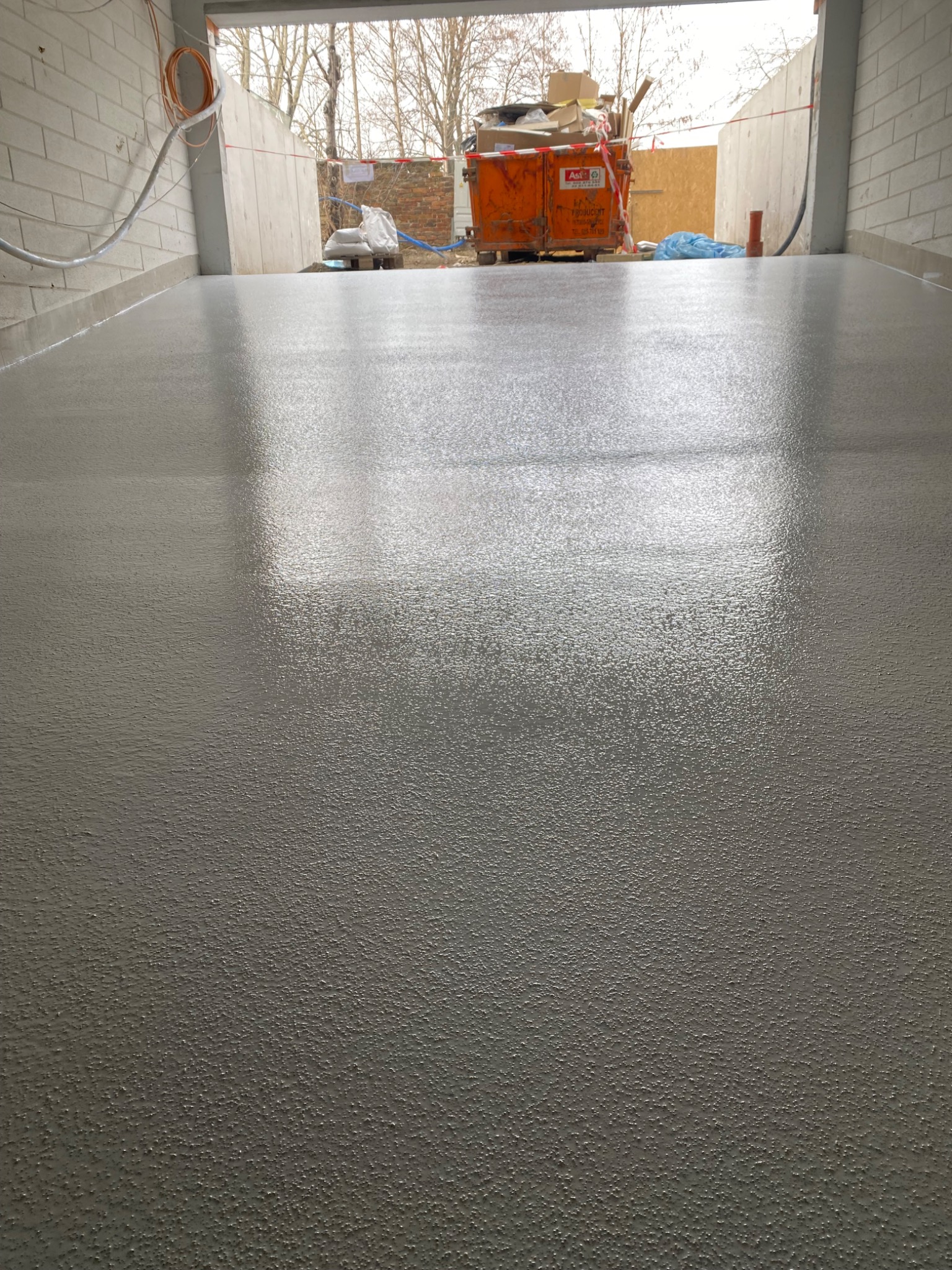 Świeżo wylana, szara posadzka w hali produkcyjnej z widocznym kontenerem na gruz w tle.