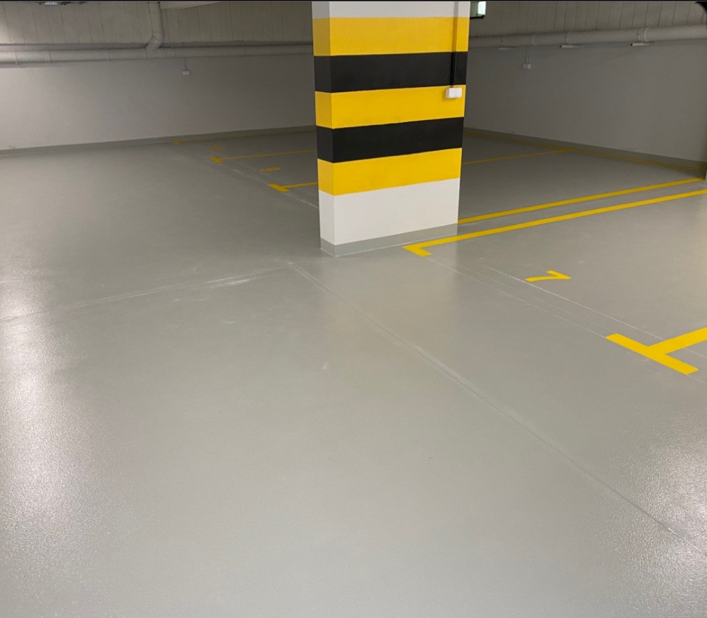 Jasnoszara, gładka posadzka w garażu podziemnym z żółtymi liniami wyznaczającymi miejsca parkingowe i numerem '1'. Kolumna w żółto-czarne pasy przy ścianie.