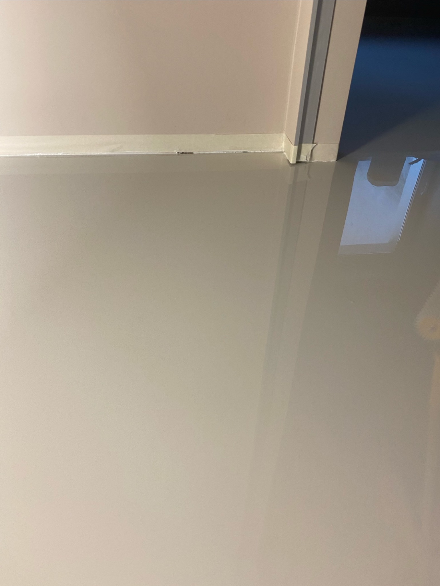 Jasnoszara, gładka posadzka z żywicy, zabezpieczona taśmą malarską przy ścianie i ościeżnicy drzwi, z widocznym odbiciem światła.