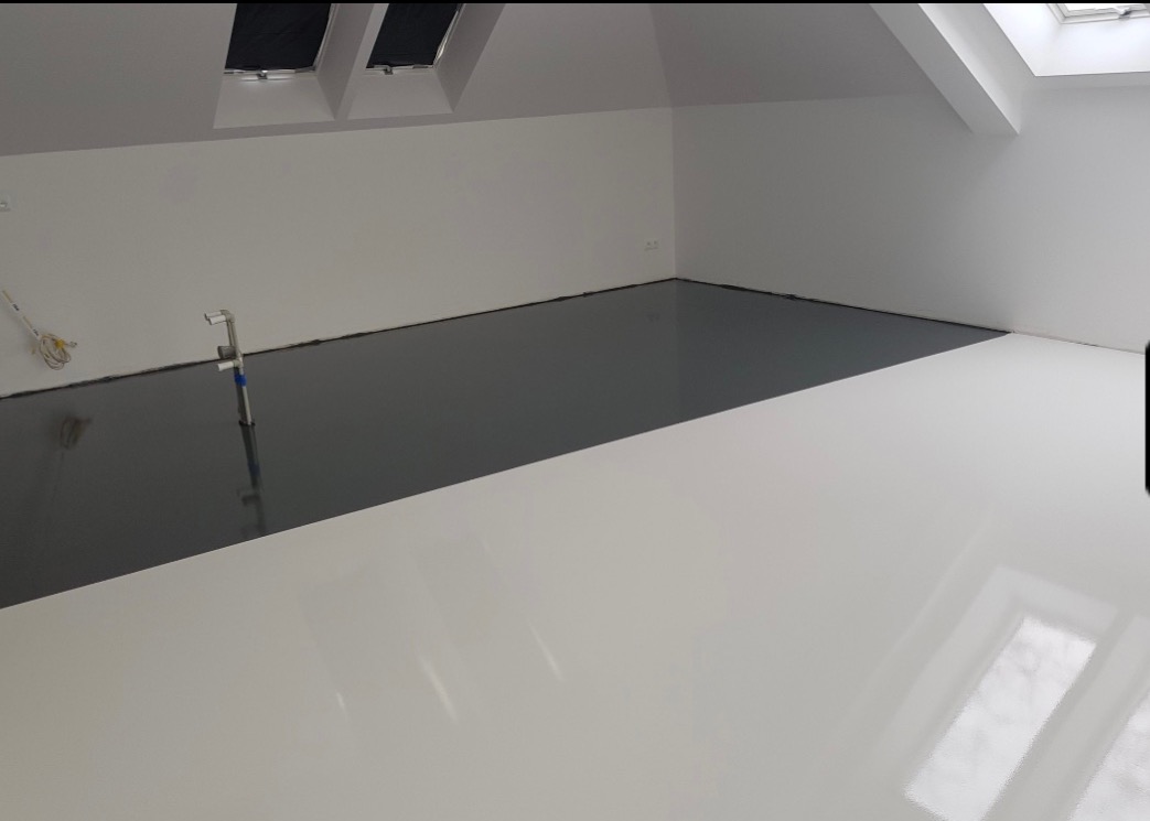 Dwukolorowa, gładka podłoga: połowa w odcieniach szarości, połowa biała, z widocznym sprzętem instalacyjnym przy ścianie i oknami dachowymi.