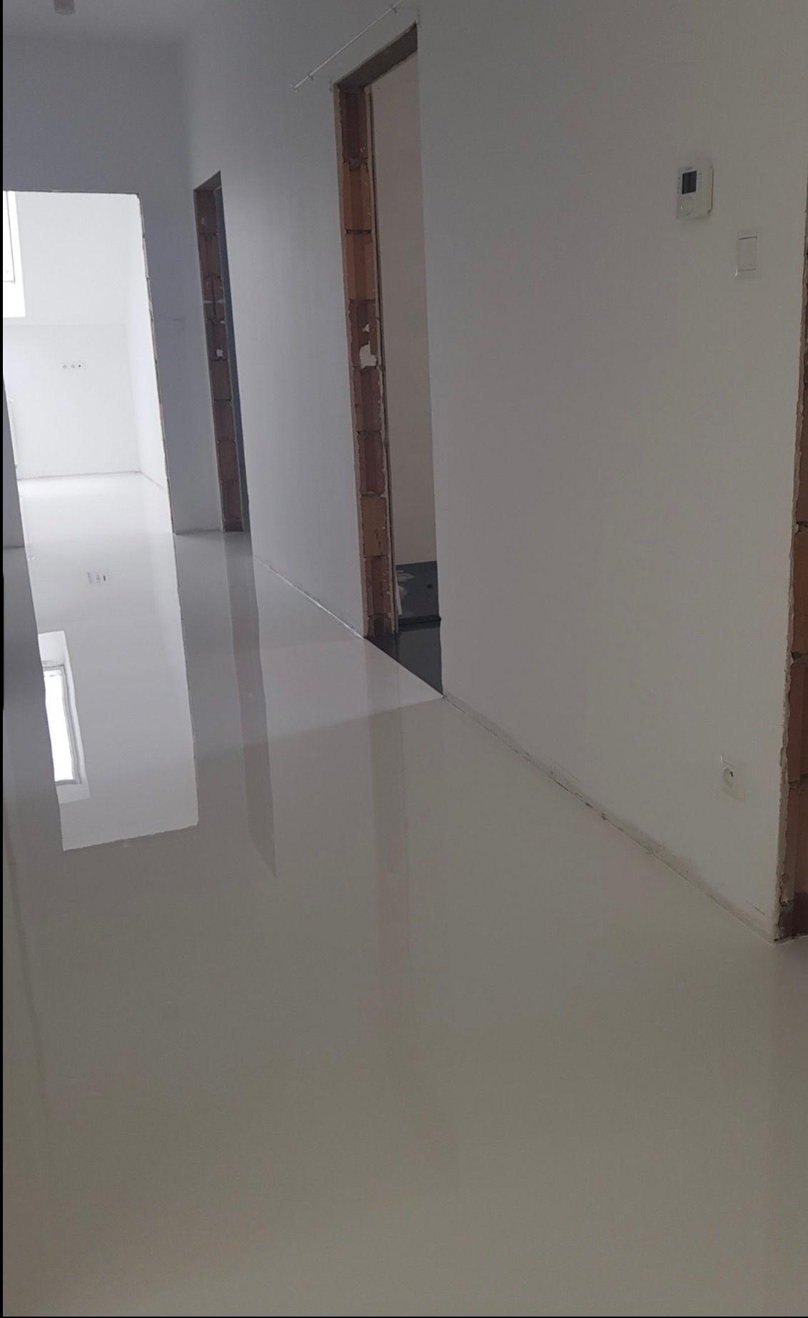 Błyszcząca, jasna podłoga w korytarzu z widocznymi drzwiami w stanie surowym i ścianami w kolorze złamanej bieli, odbijająca światło okien i lamp, z widocznym termostatem na ścianie.