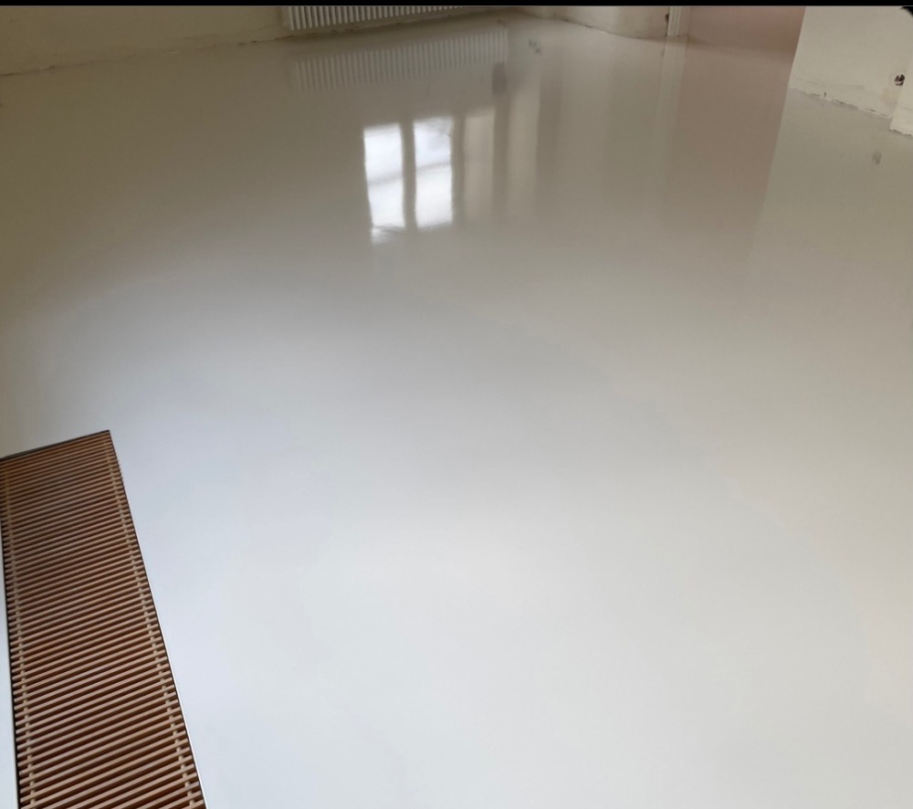 Jasna, jednolita podłoga z połyskiem, prawdopodobnie żywiczna, z widocznym odbiciem okna i kratką wentylacyjną w rogu