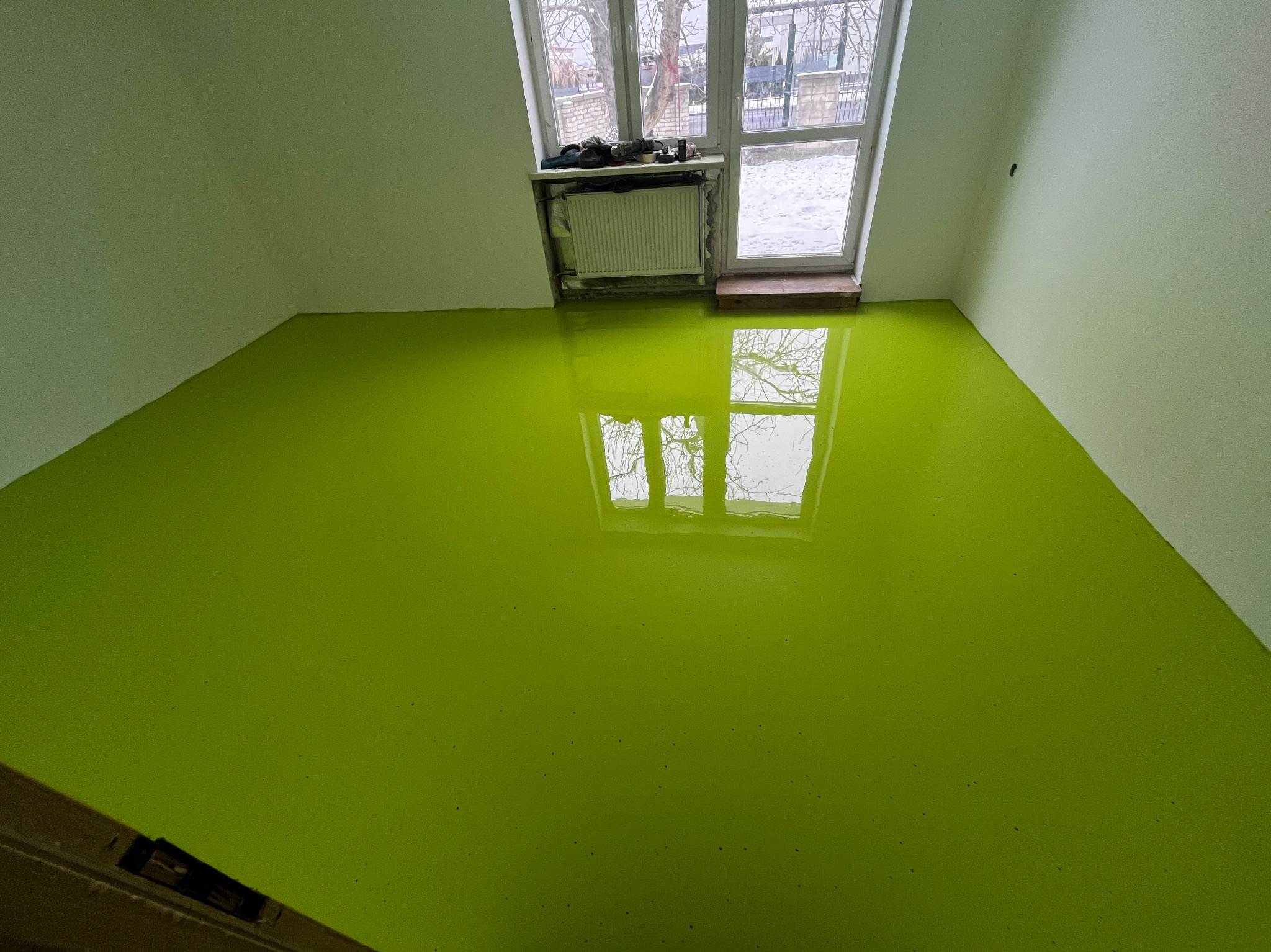 Świeżo wylana, jaskrawo zielona posadzka w pokoju w Warszawie, z odbiciem okna w gładkiej powierzchni.