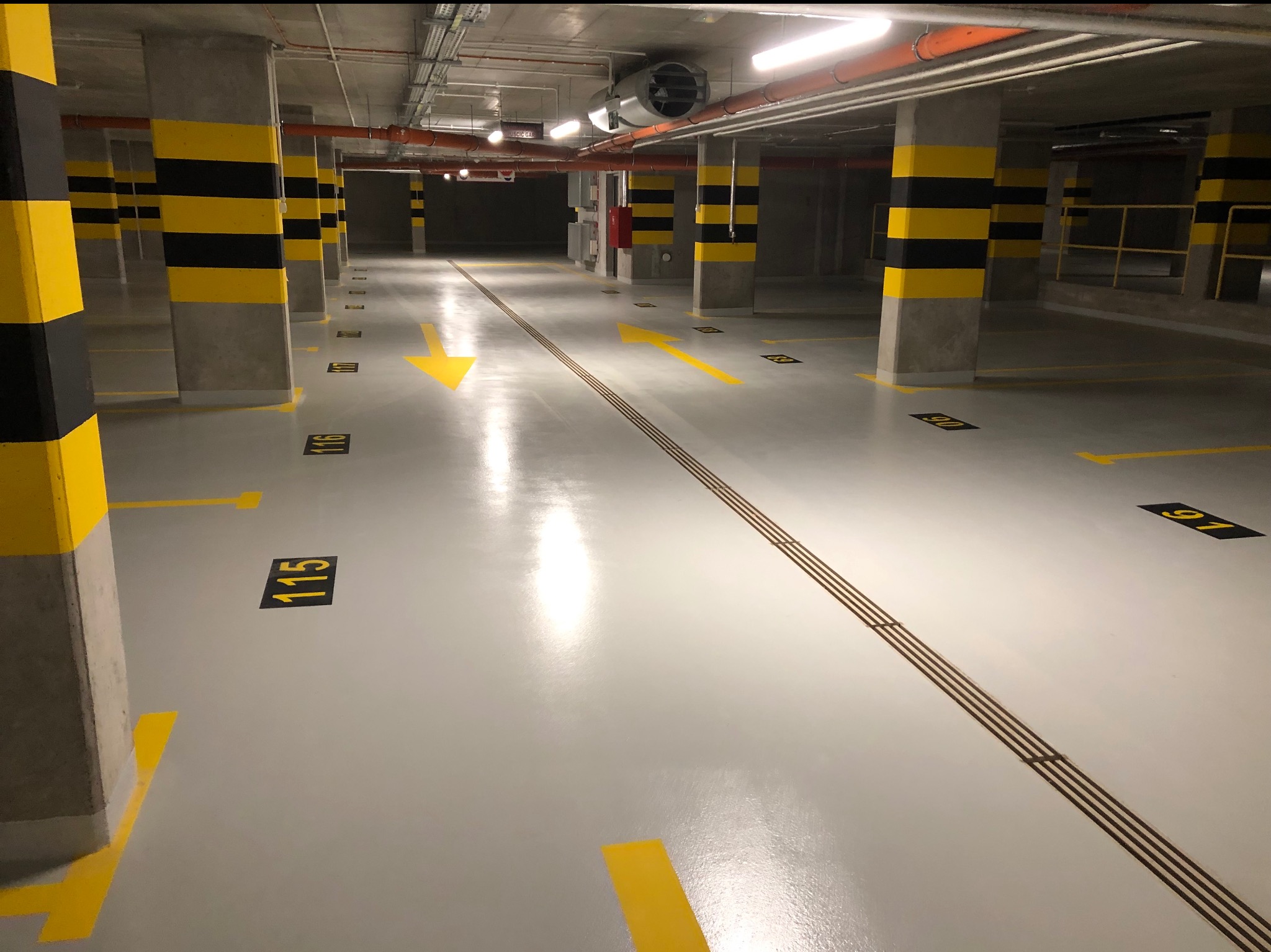 Biała, błyszcząca posadzka w garażu podziemnym z żółtymi oznaczeniami stanowisk, numerami i strzałkami kierunkowymi; kolumny pomalowane w czarno-żółte pasy.