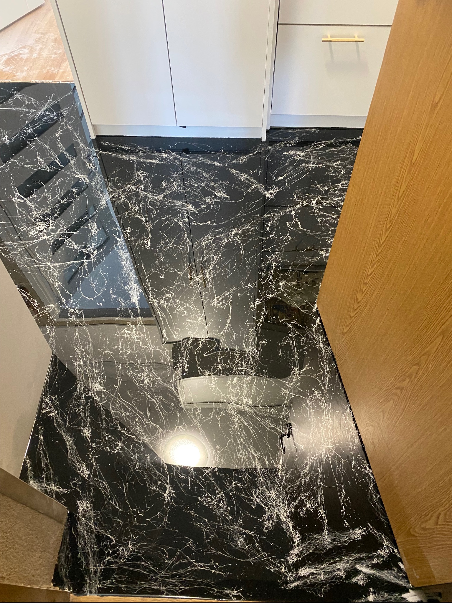 Czarna posadzka z białymi żyłkami, przypominająca marmur, sąsiaduje z białymi szafkami i drewnianą ścianą. Widok z góry, połyskująca powierzchnia.