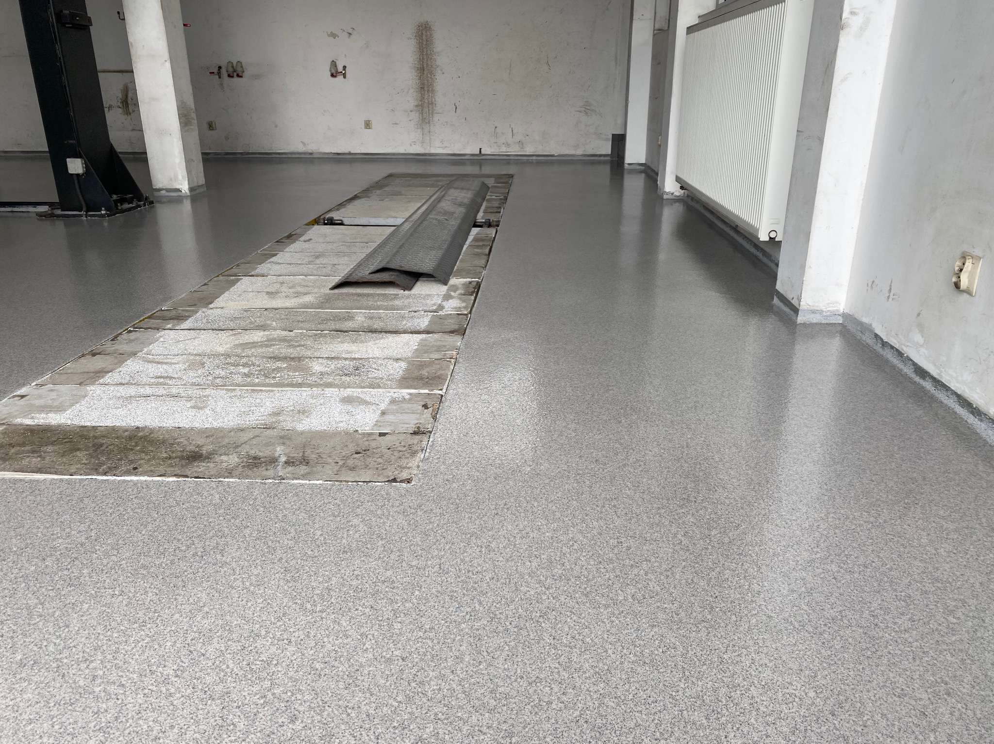 Szara, gładka posadzka z drobnym wzorem w przemysłowym wnętrzu. Widoczny kanał rewizyjny i fragmenty instalacji na ścianach. Nowoczesne wykończenie podłogi.