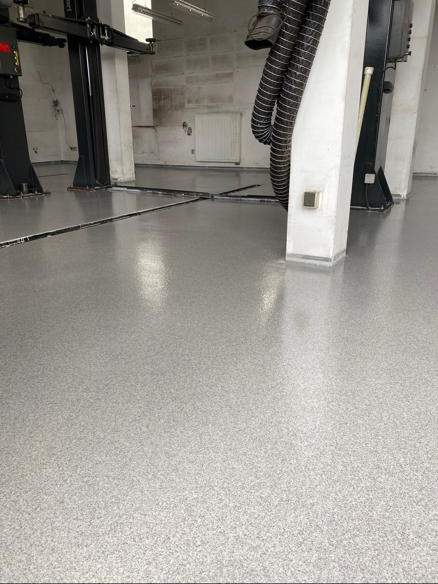 Błyszcząca, jasnoszara posadzka w hali warsztatowej z podnośnikami samochodowymi. Nowa, gładka powierzchnia, z widocznymi szczelinami dylatacyjnymi.