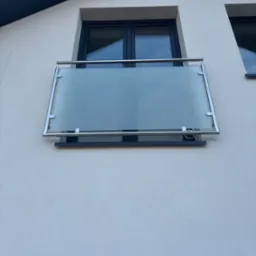 Szklana barierka balkonowa z nierdzewnymi elementami mocującymi, zamontowana na fasadzie budynku z ciemnymi oknami.