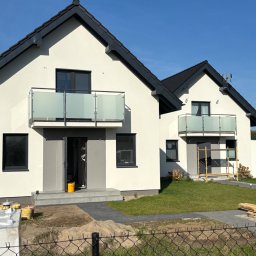 MT BALUSTRADY Maciej Kamiński - Dwa nowoczesne domy jednorodzinne z balkonami zabezpieczonymi matowym szkłem i metalową ramą, widoczne elementy wykończeniowe wokół posesji.