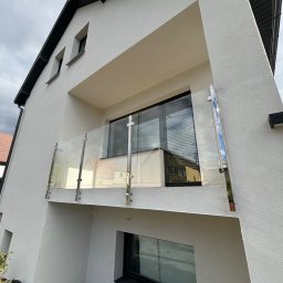 MT BALUSTRADY Maciej Kamiński - Nowoczesny balkon z przeszkloną barierą na tle białej elewacji domu z ciemną stolarką okienną, widoczne materiały budowlane na posadzce.