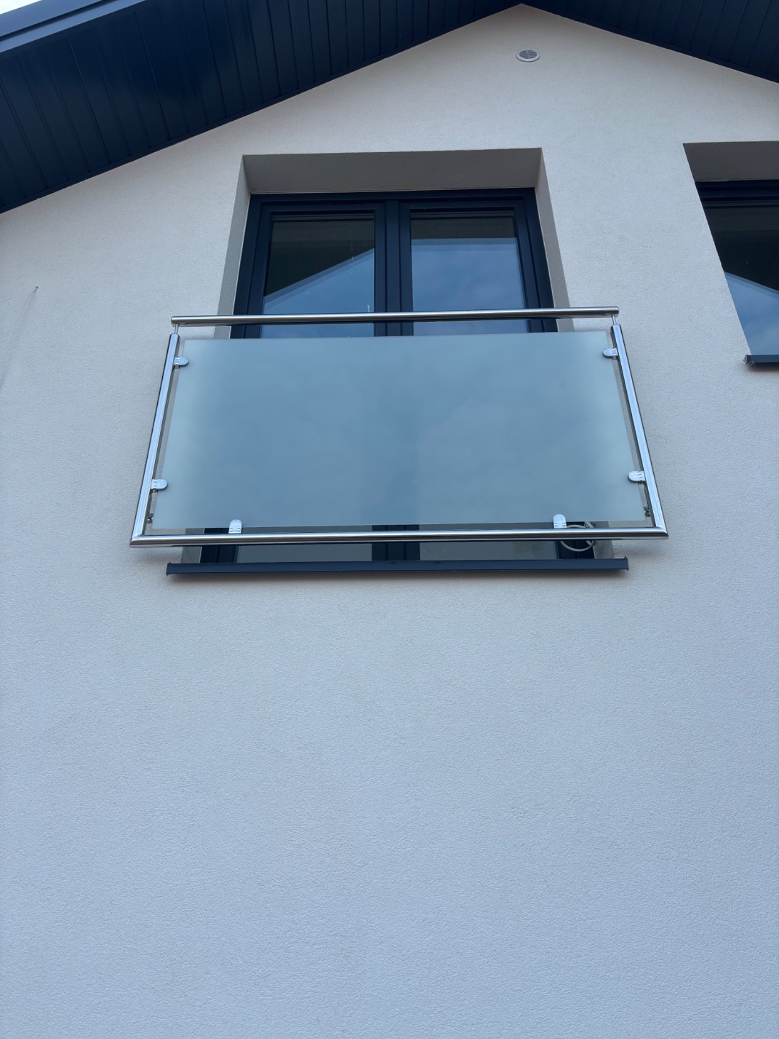 Szklana barierka balkonowa z nierdzewnymi elementami mocującymi, zamontowana na fasadzie budynku z ciemnymi oknami.