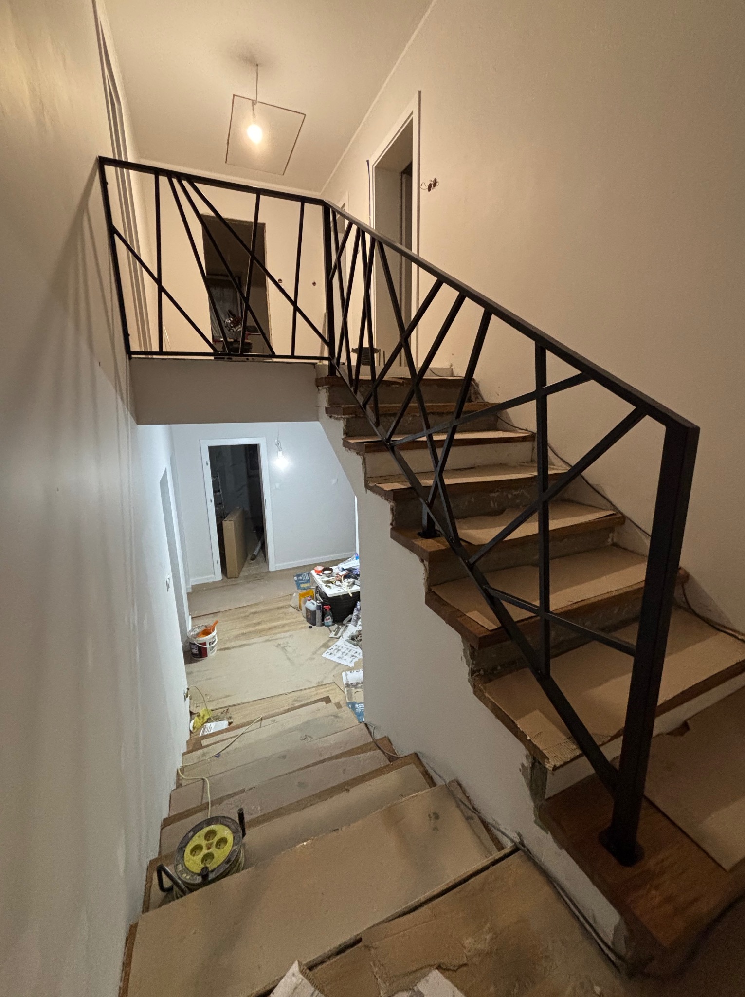 Schody w trakcie remontu z zamontowaną czarną, geometryczną, metalową konstrukcją zabezpieczającą, widoczny zwój kabla elektrycznego na jednym ze stopni.