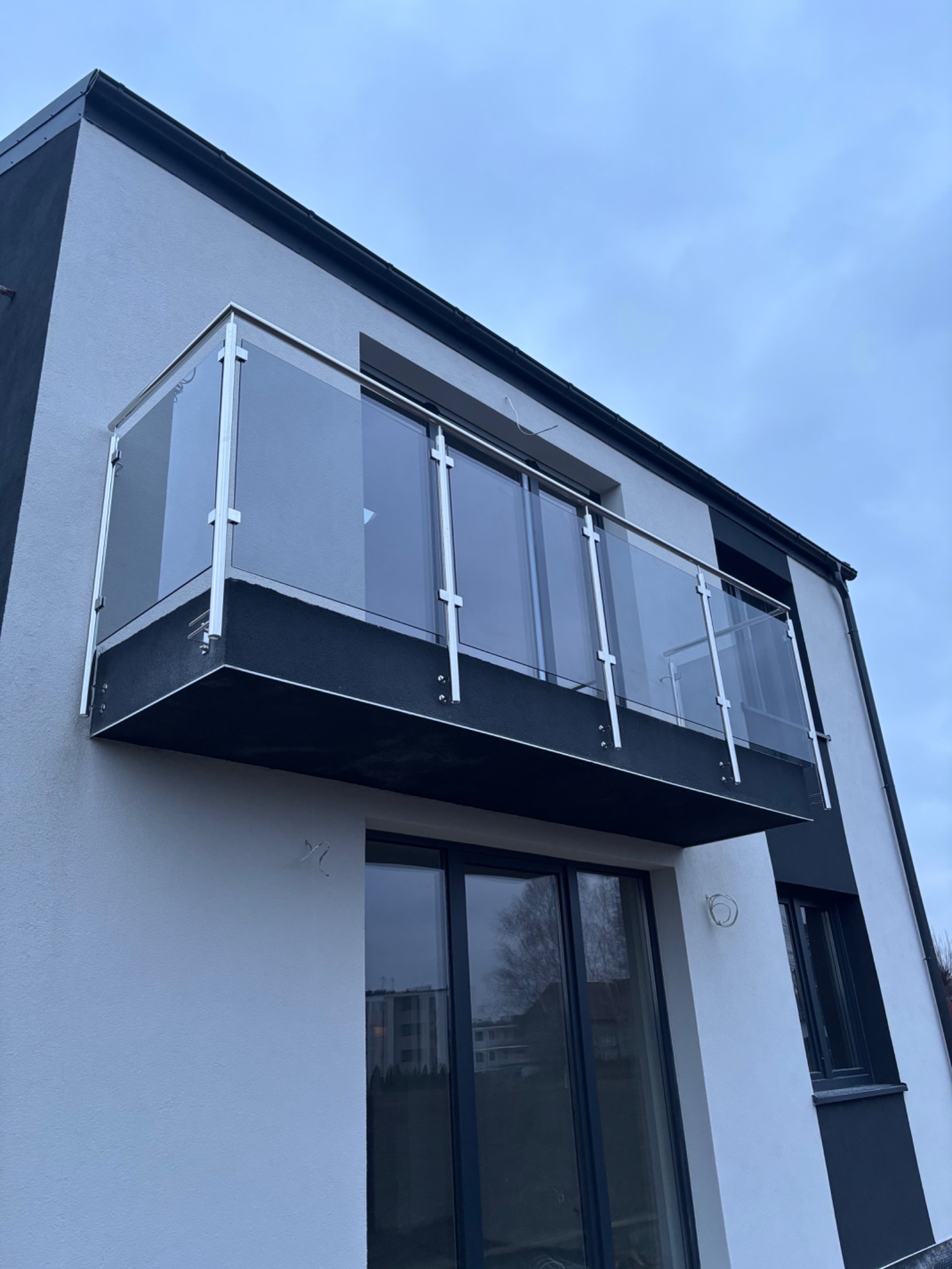 Nowoczesny balkon z przezroczystą szklaną taflą i metalowymi słupkami, widok z dołu na tle pochmurnego nieba.