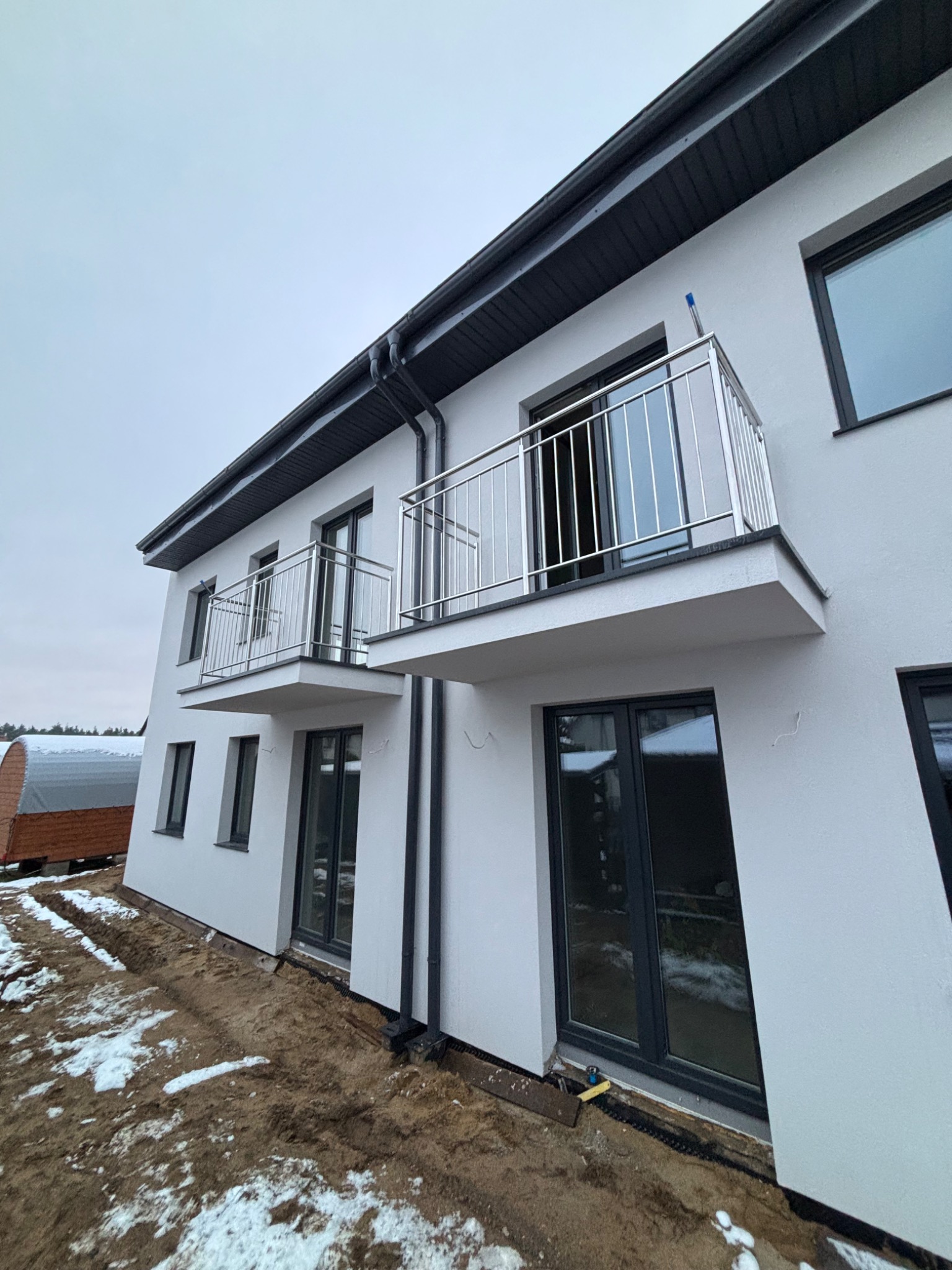 Nowoczesny dom z dwoma balkonami z balustradami nierdzewnymi, widok z dołu na elewację i częściowo zagospodarowany teren.