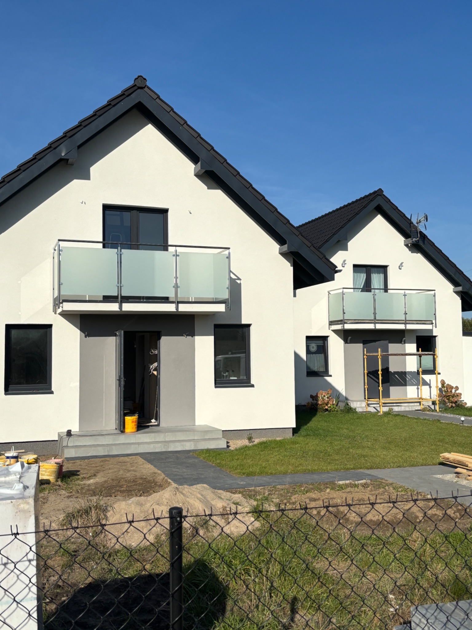 Dwa nowoczesne domy jednorodzinne z balkonami zabezpieczonymi matowym szkłem i metalową ramą, widoczne elementy wykończeniowe wokół posesji.