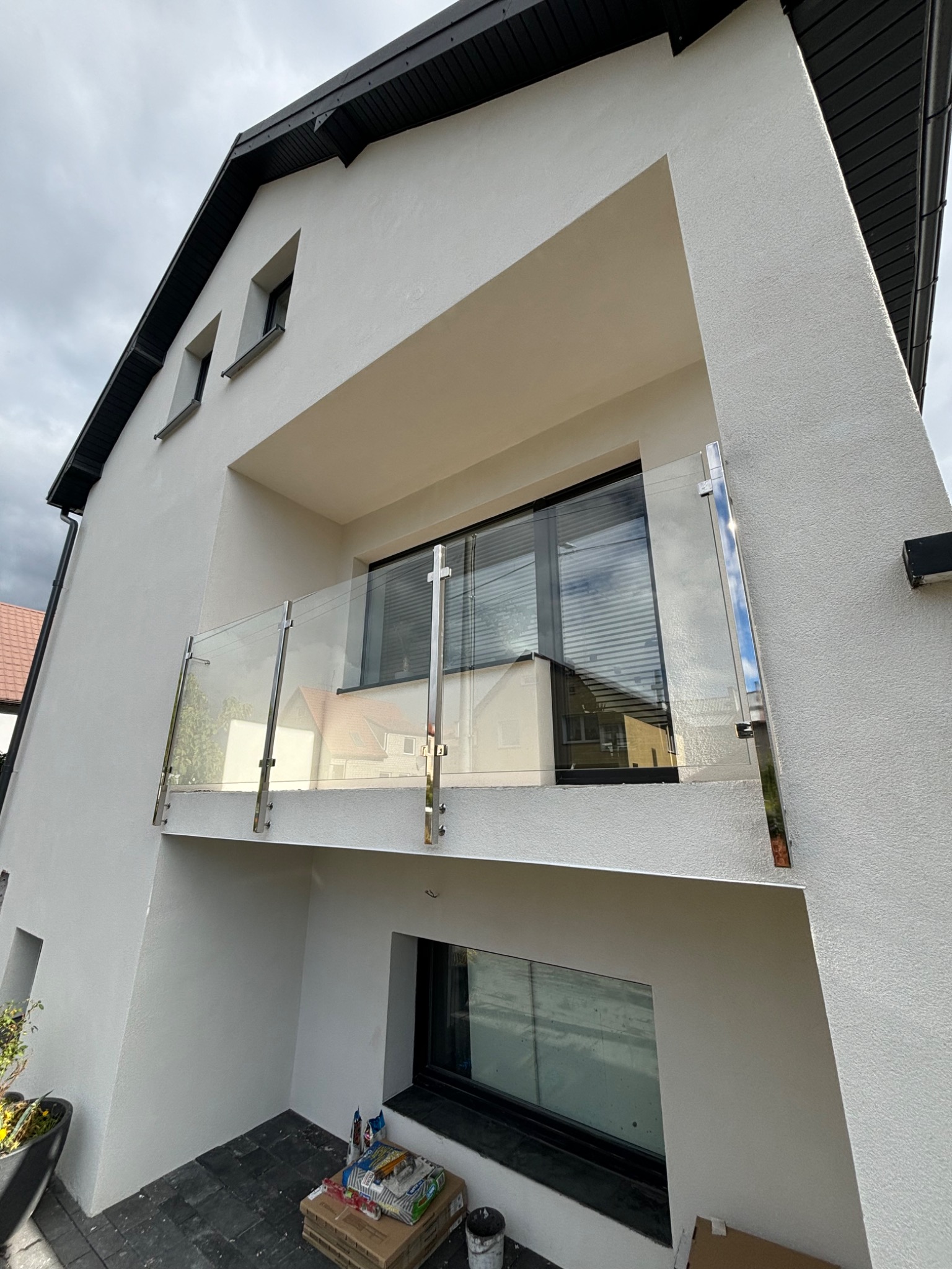 Nowoczesny balkon z przeszkloną barierą na tle białej elewacji domu z ciemną stolarką okienną, widoczne materiały budowlane na posadzce.