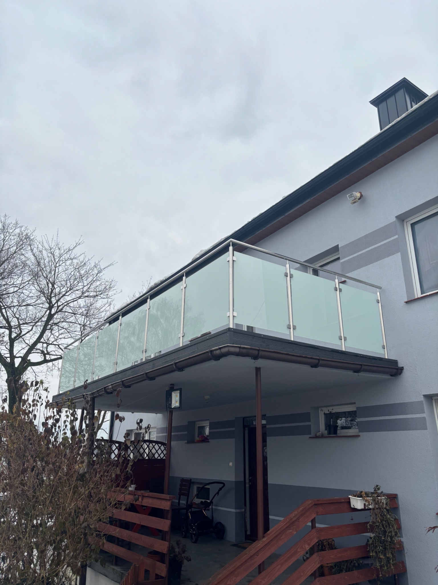 Zewnętrzny balkon z nowoczesną szklaną balustradą i nierdzewnymi słupkami, widok z dołu, na tle pochmurnego nieba i elewacji budynku z drewnianymi elementami wykończeniowymi.