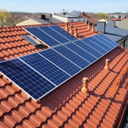 PIEDOX Piotr Pałka - Panele fotowoltaiczne zamontowane na dachu z czerwonej dachówki ceramicznej z kominkami wentylacyjnymi; widoczne okna dachowe i fragmenty innych budynków w tle.