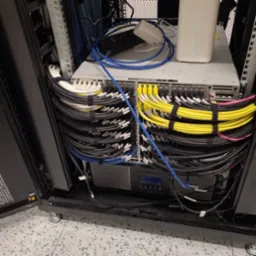 Zamknięta szafa rack z uporządkowanymi kablami sieciowymi oznaczonymi etykietami, widoczne switche, router, listwa zasilająca i okablowanie w kolorach białym, żółtym, niebieskim i czarnym, spięte...