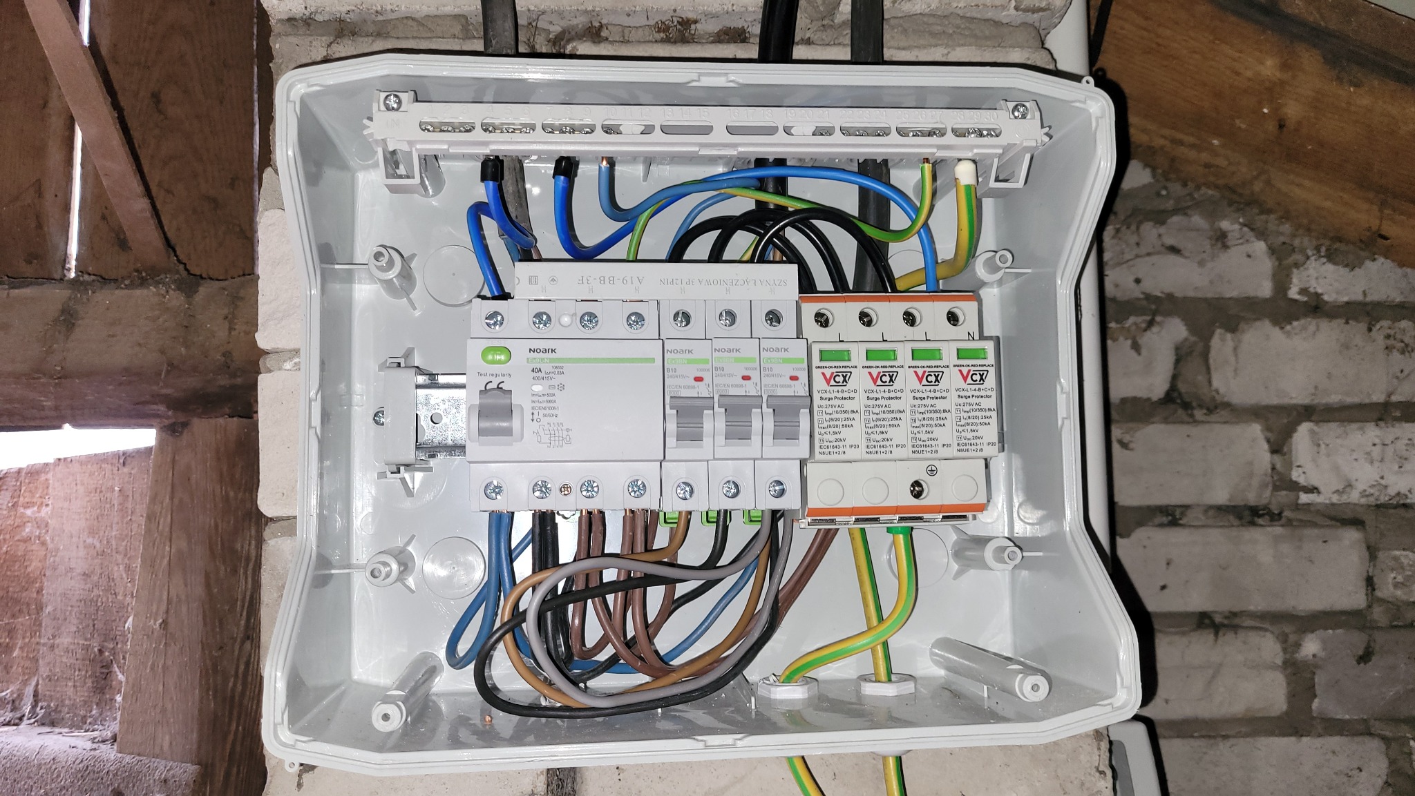Otwarta skrzynka elektryczna z zamontowanymi bezpiecznikami, ogranicznikami przepięć i podłączonymi przewodami o różnych kolorach, zamontowana na ceglanej ścianie w surowym wnętrzu.