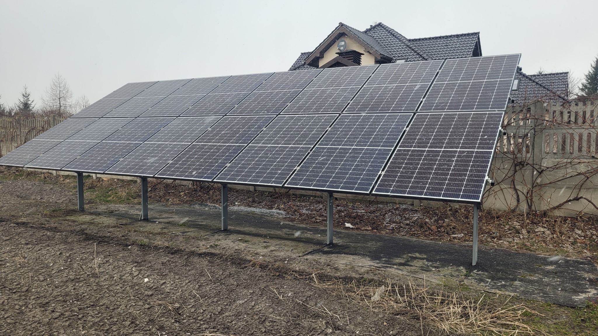 Panel słoneczny na metalowych wspornikach, ustawiony przed domem z widocznym fragmentem dachu i komina, w tle szary betonowy płot z suchymi pnączami, pochmurny dzień.