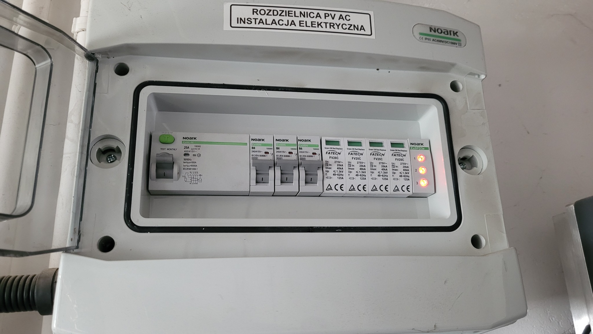 Otwarta skrzynka rozdzielcza PV AC z zainstalowanymi zabezpieczeniami przepięciowymi i wyłącznikami nadprądowymi marki Noark i Fatech, widoczne diody sygnalizacyjne stanu.