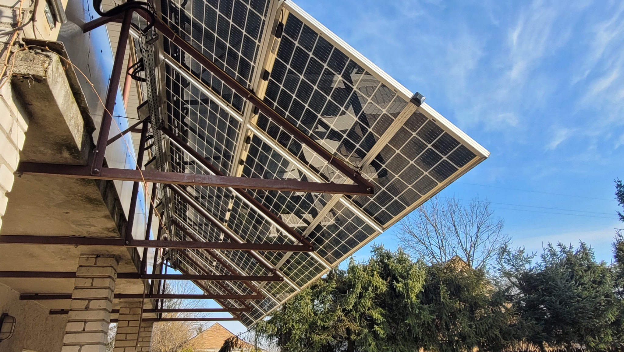 Panele fotowoltaiczne zamontowane jako zadaszenie nad wejściem, widoczne z dołu pod kątem, z metalową konstrukcją wsporczą i fragmentem muru budynku.