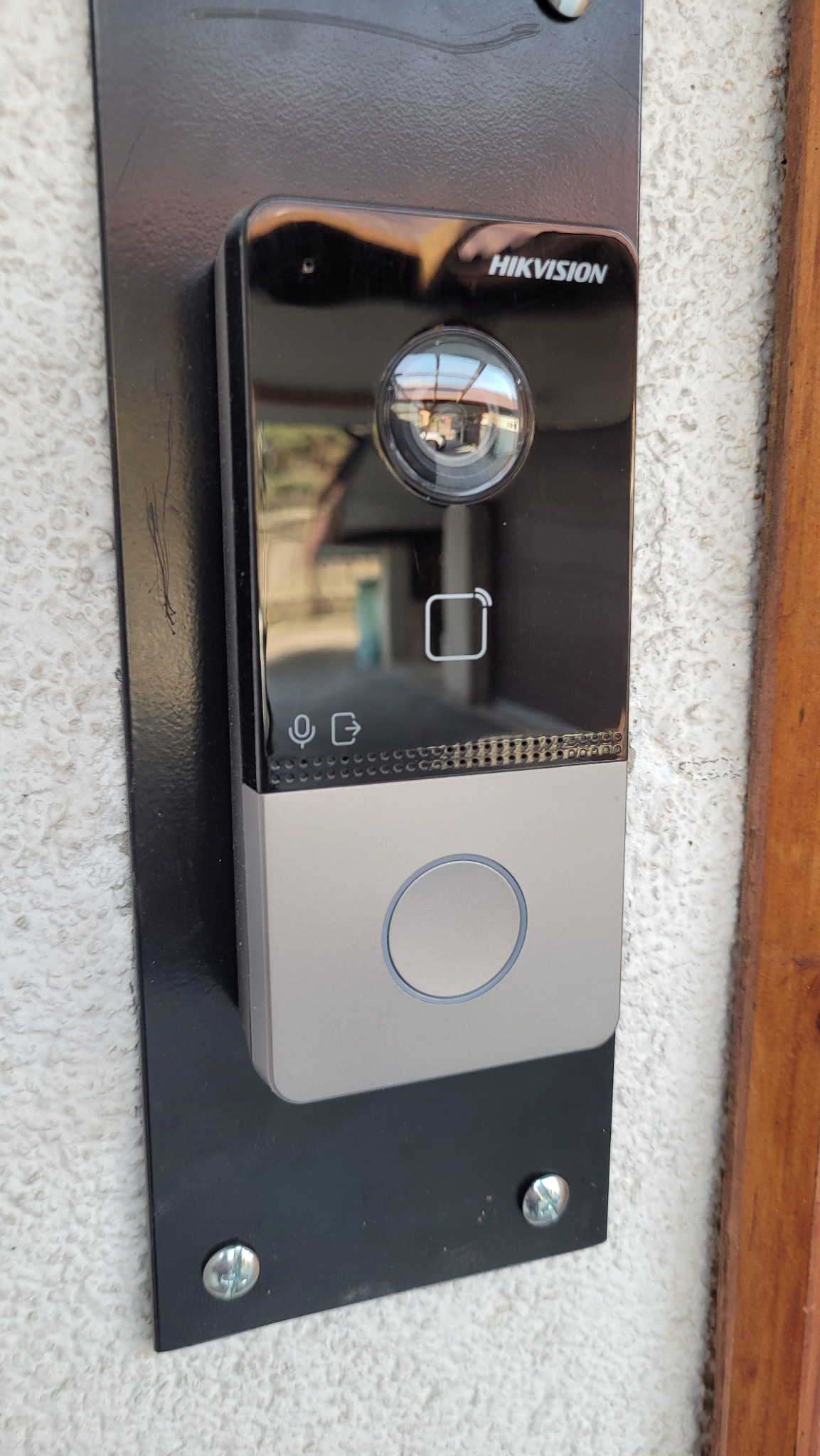 Zewnętrzny panel domofonowy Hikvision z kamerą, czytnikiem kart i przyciskiem dzwonka, zamontowany na czarnej blasze przykręconej do ściany o nierównej fakturze.