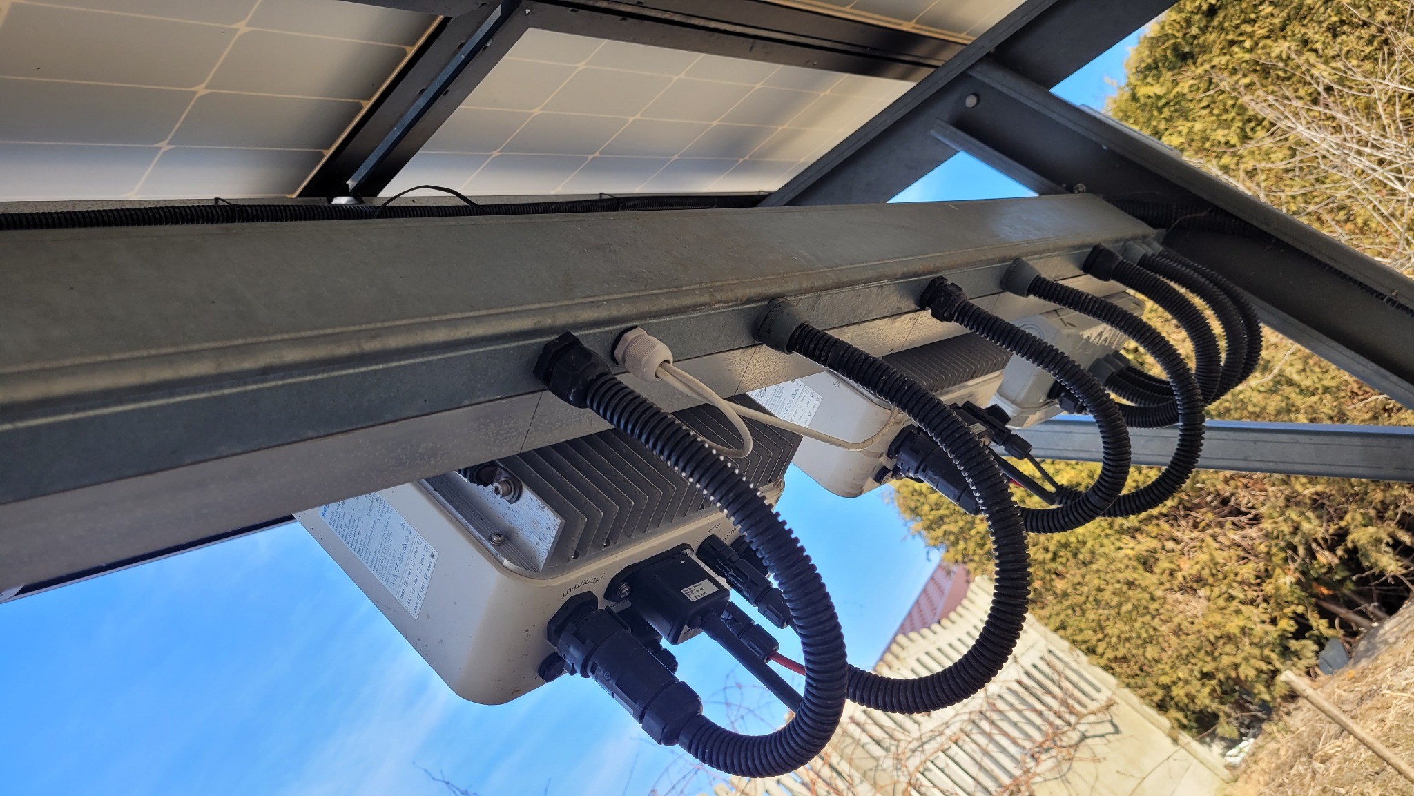 Widok od dołu na zamontowane panele słoneczne z widocznymi inwerterami i okablowaniem w czarnych peszlach, na tle błękitnego nieba i drzew.