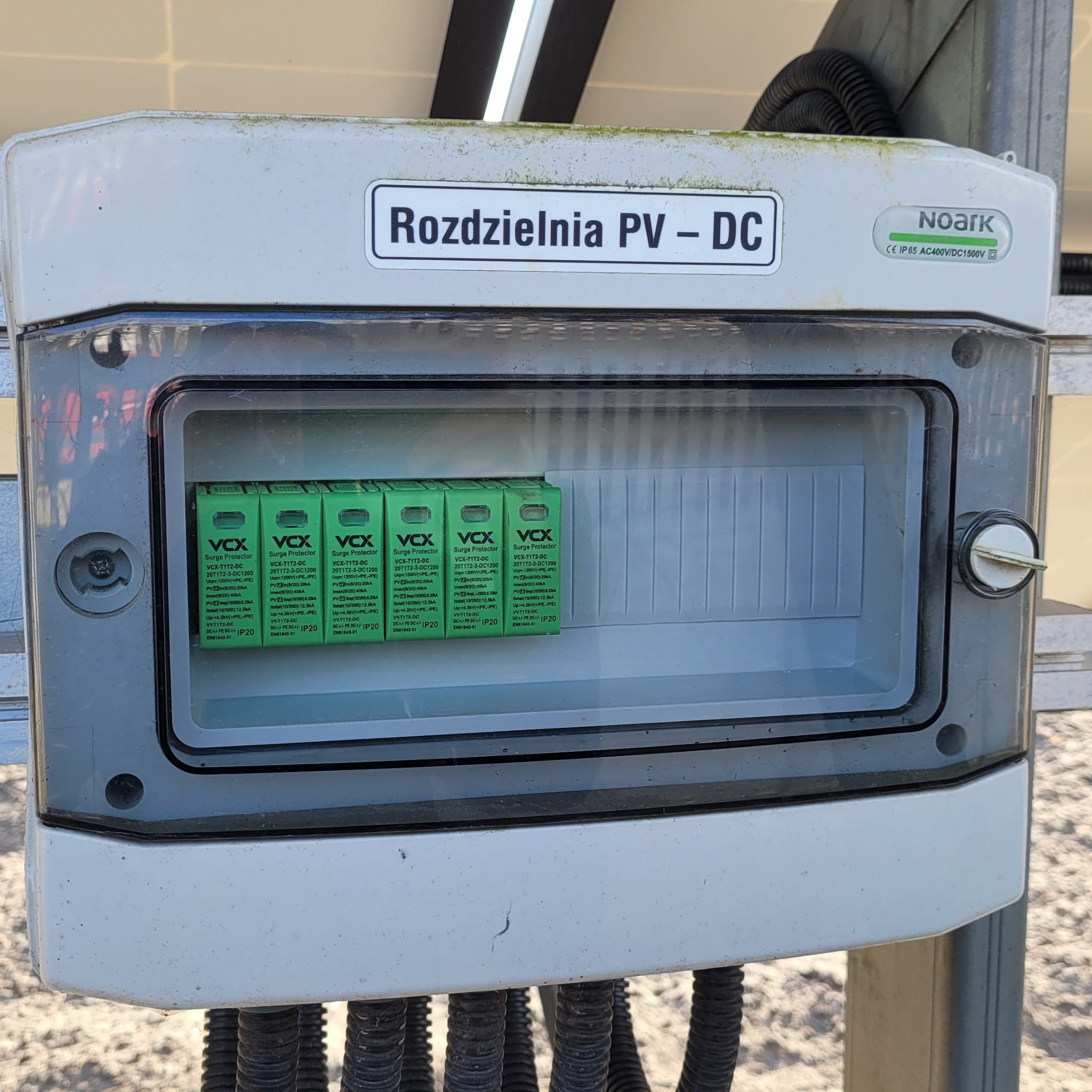 Rozdzielnia PV-DC marki Noark z widocznymi modułowymi ogranicznikami przepięć VCX wewnątrz, zamontowana na konstrukcji paneli fotowoltaicznych.