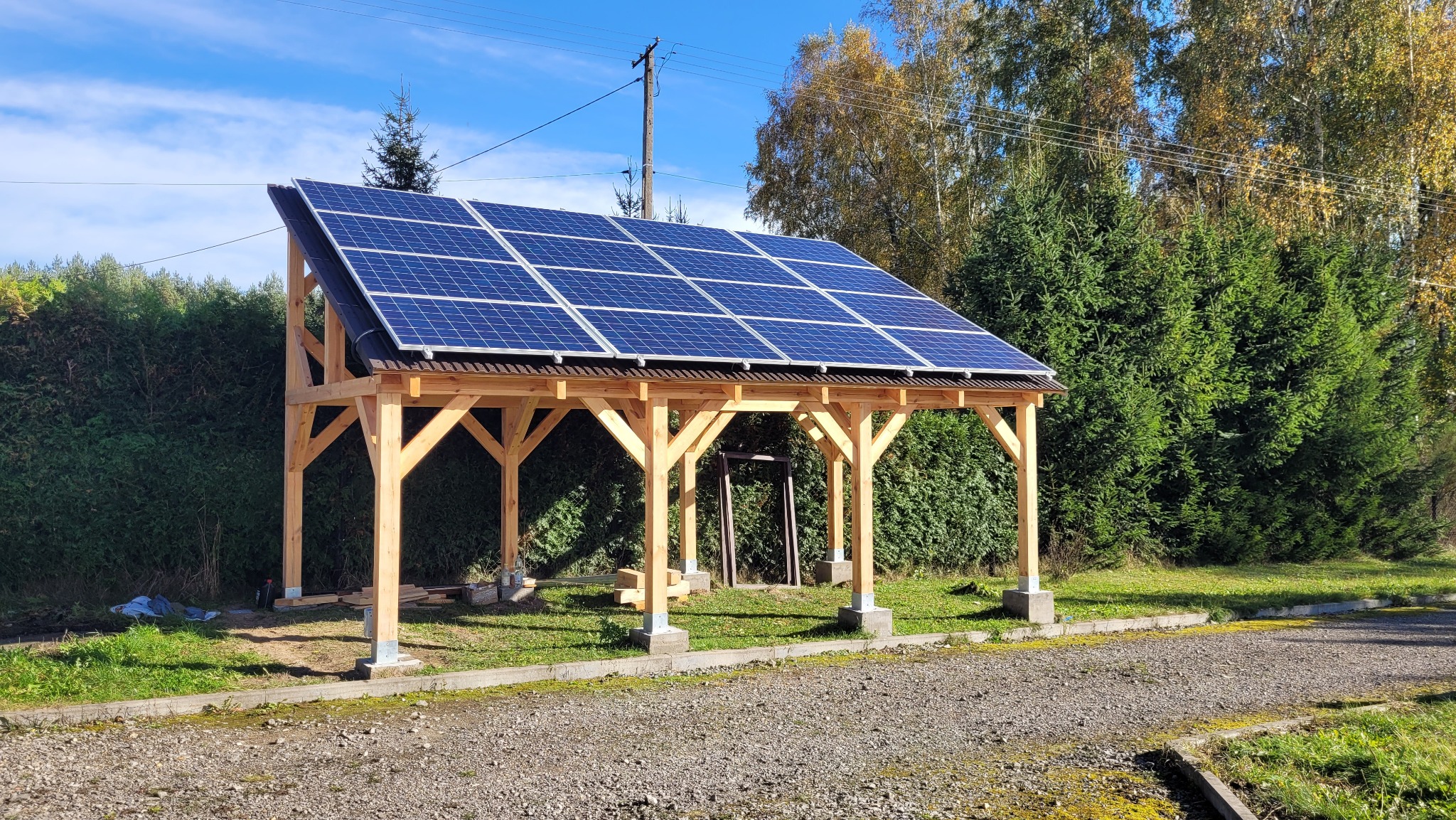 Drewniana wiata z panelami słonecznymi na dachu, widoczna w słoneczny dzień na tle zieleni.