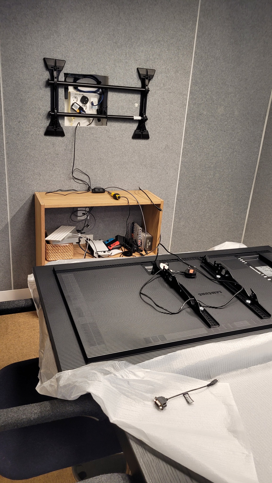 Przygotowanie do montażu telewizora: uchwyt ścienny zamontowany, telewizor Samsung leży na stole z przykręconymi mocowaniami, obok drewniany regał z akcesoriami i narzędziami.