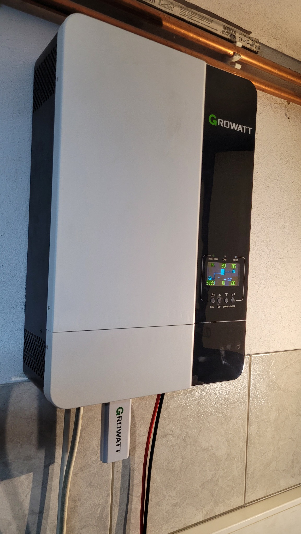 Biało-czarny inwerter Growatt zamontowany na ścianie z beżowych płytek, widoczne przewody, miedziana rurka i wyświetlacz pokazujący dane.